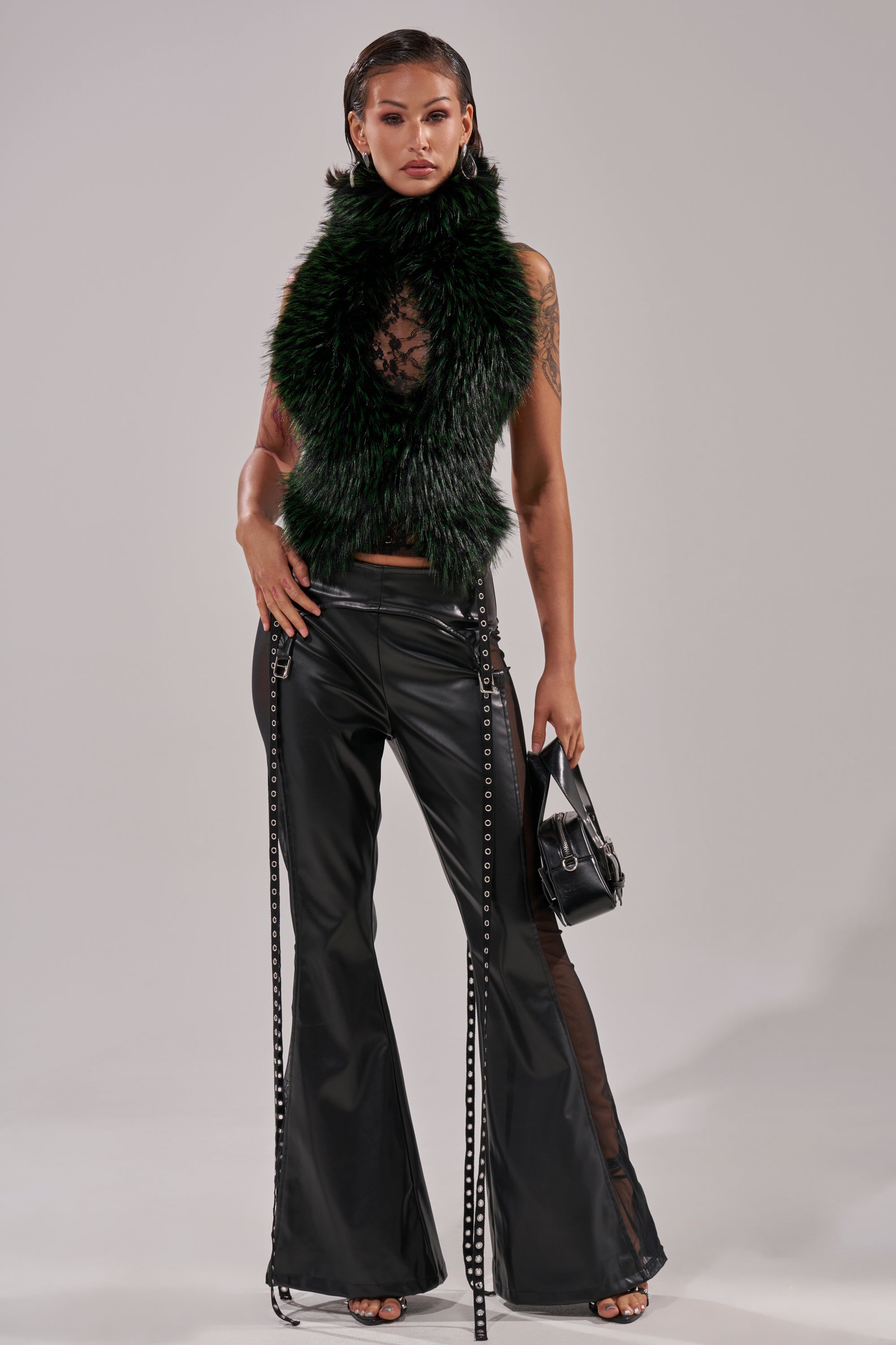 Confidently posing against a plain light background, a person wears the FEEL ME BABY FUR BLOUSE, sheer black top, black lace-up leather pants, and heels, accessorized with a small black handbag.