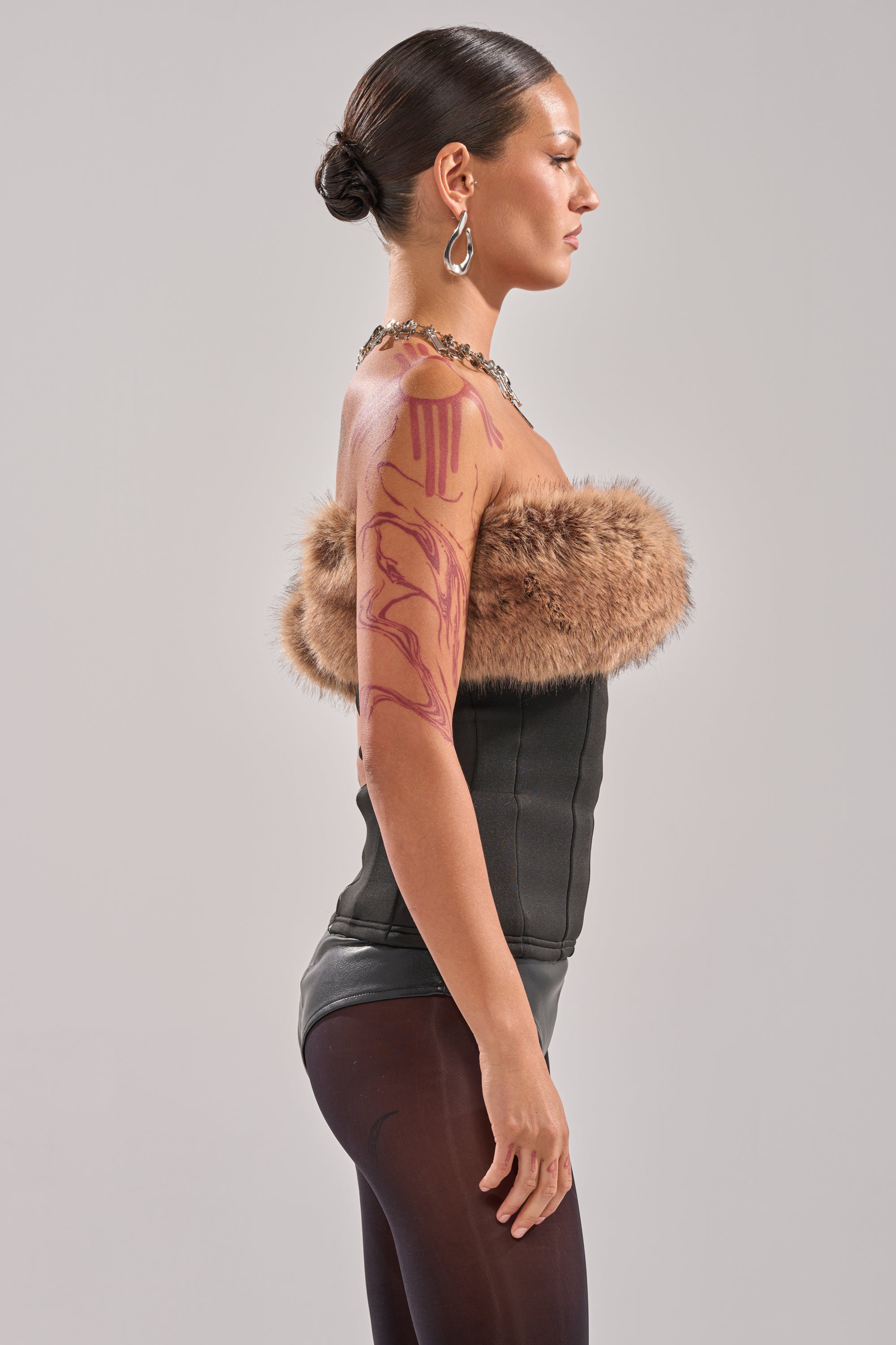 A woman in profile poses against a plain background wearing the SO CHIC FAUX LEATHER BLOUSE WITH FUR TRIM, black pants, large hoop earrings, and a necklace. Her hair is in a bun and bold pink body art adorns her arm. 6