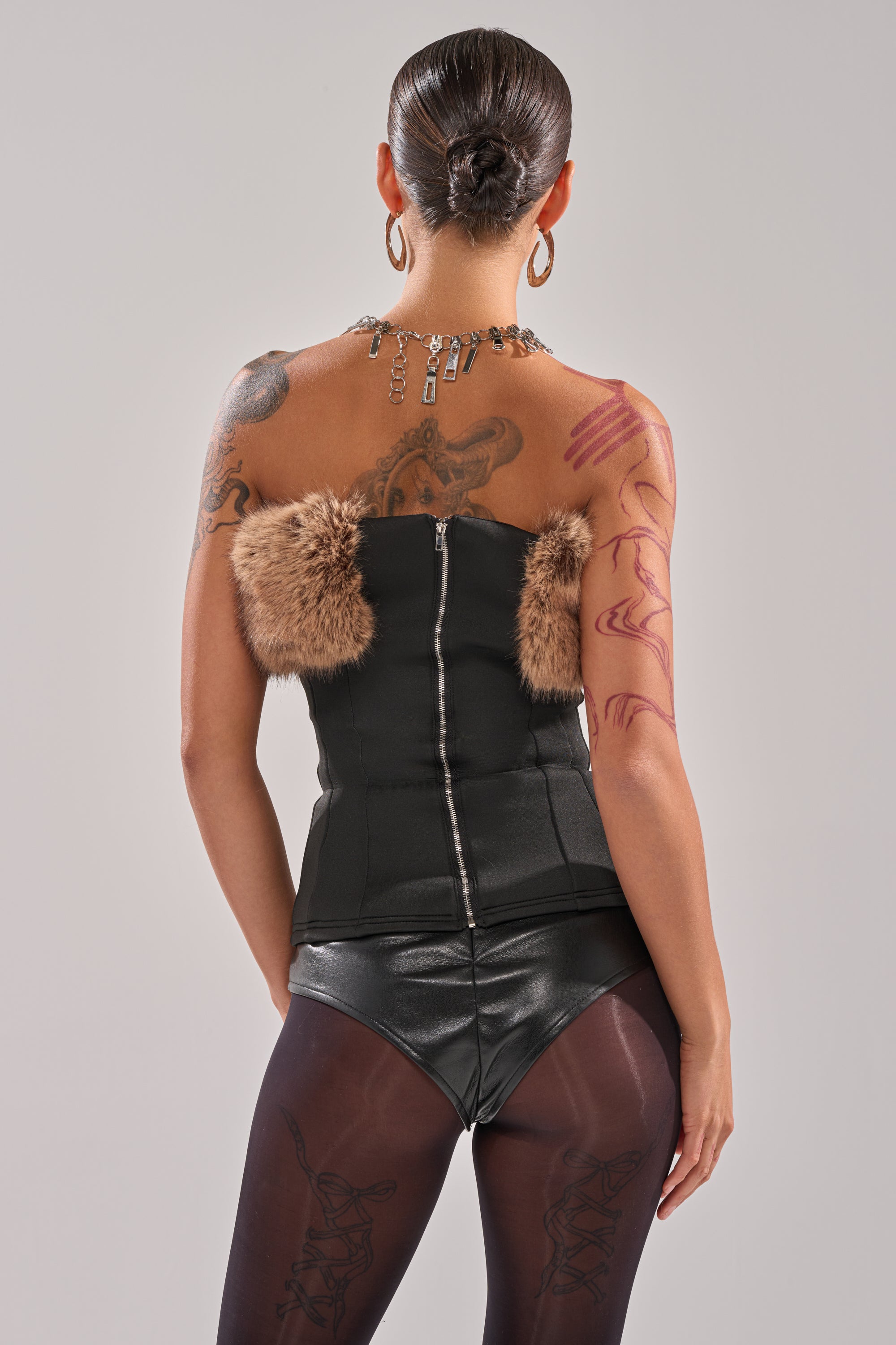 A person with tattoos and short hair faces away, wearing the SO CHIC FAUX LEATHER BLOUSE WITH FUR TRIM, black shorts, sheer tights, hoop earrings, and a charm necklace against a neutral background. 7