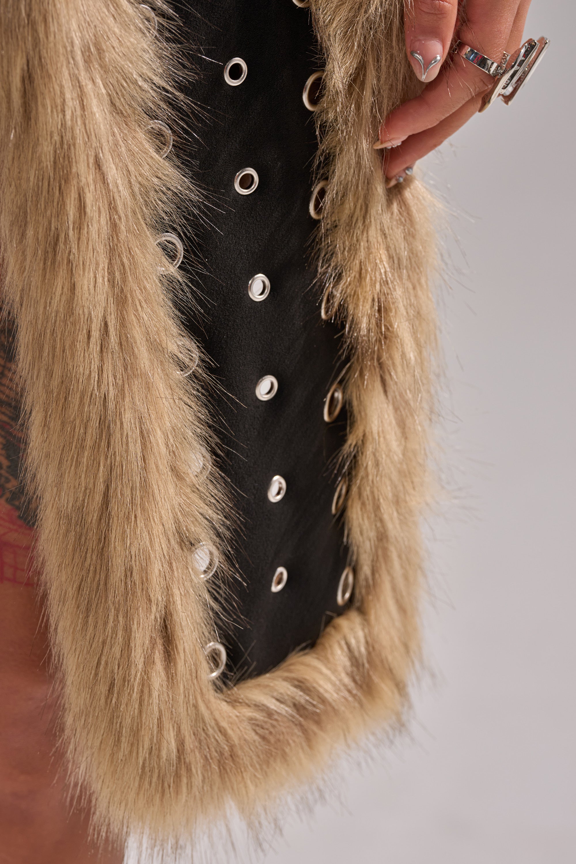 A close-up of a person wearing the ICY HOT FUR BRALETTE, featuring black fabric with metal eyelets and beige faux fur trim, with their hand adorned in rings and manicured nails visible. 9