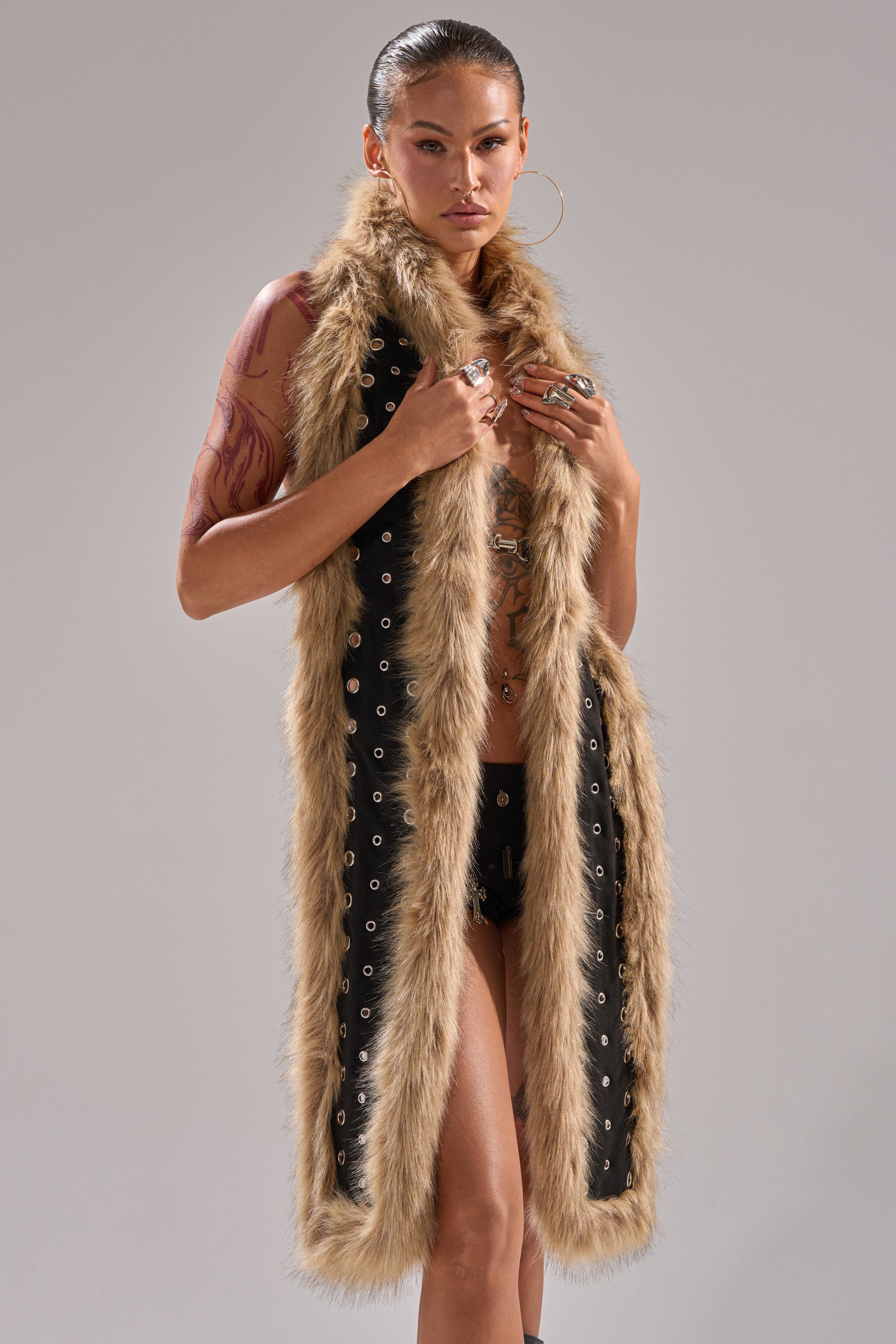 A person with slicked-back hair and hoop earrings models the ICY HOT FUR BRALETTE with tattoos and multiple rings, styled over minimal clothing, standing against a plain background. 1