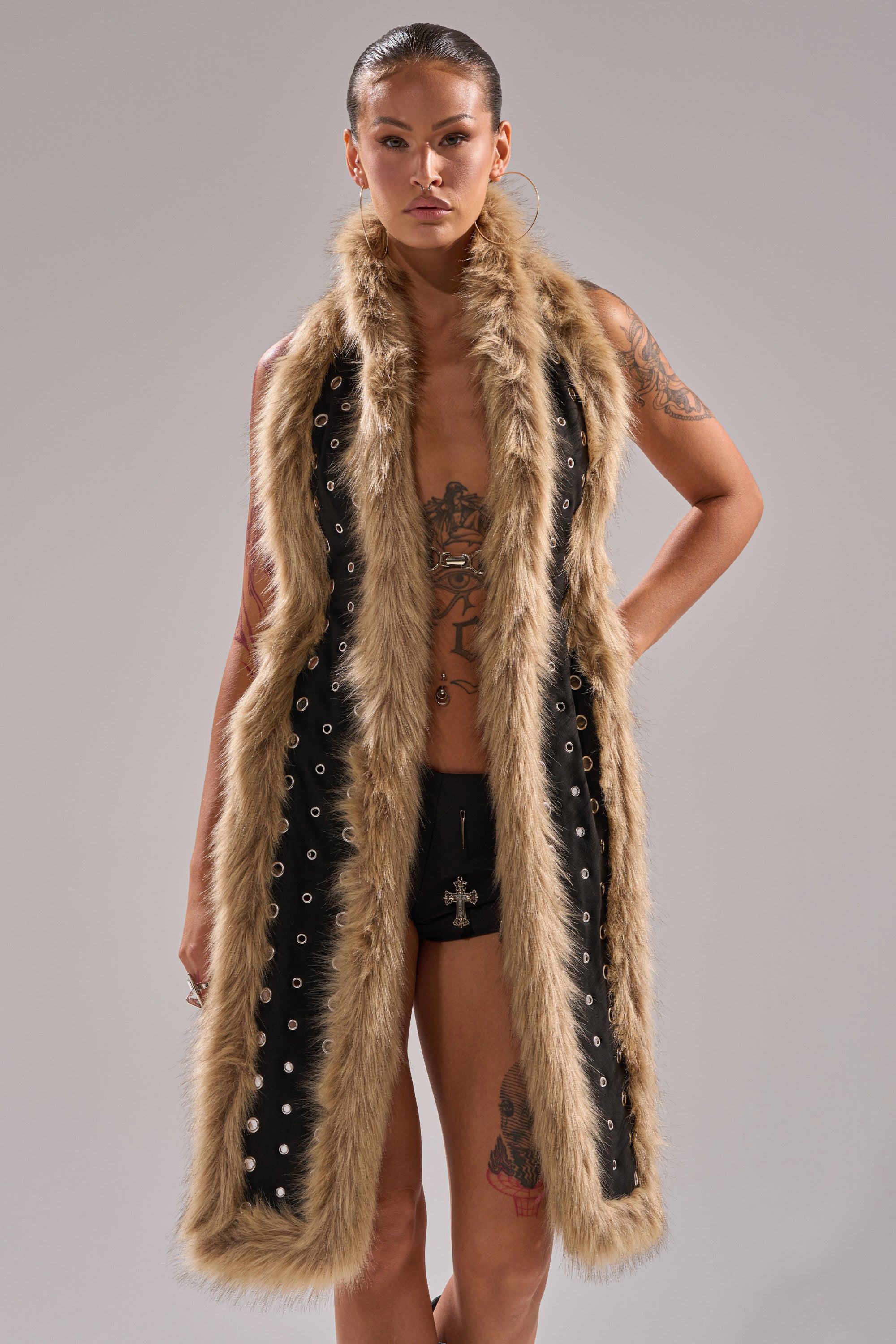 A person with tattoos and hoop earrings models the ICY HOT FUR BRALETTE—a long, sleeveless black vest with tan faux fur trim and grommets—over black shorts against a plain gray background. 2