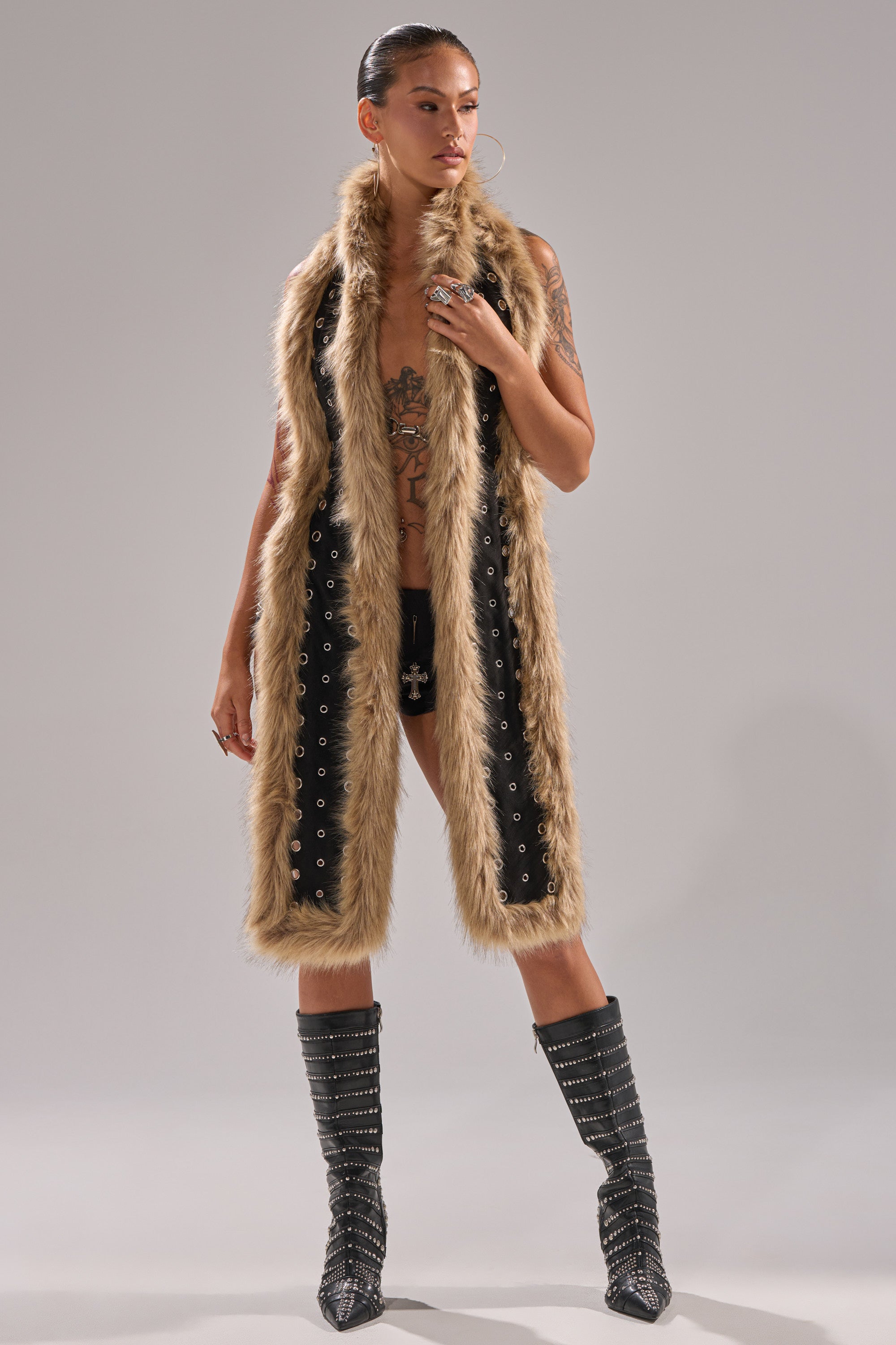 A person poses confidently in the ICY HOT FUR BRALETTE, styled with black shorts featuring a cross detail, tall studded boots, hoop earrings, and visible tattoos, set against a plain background. 7