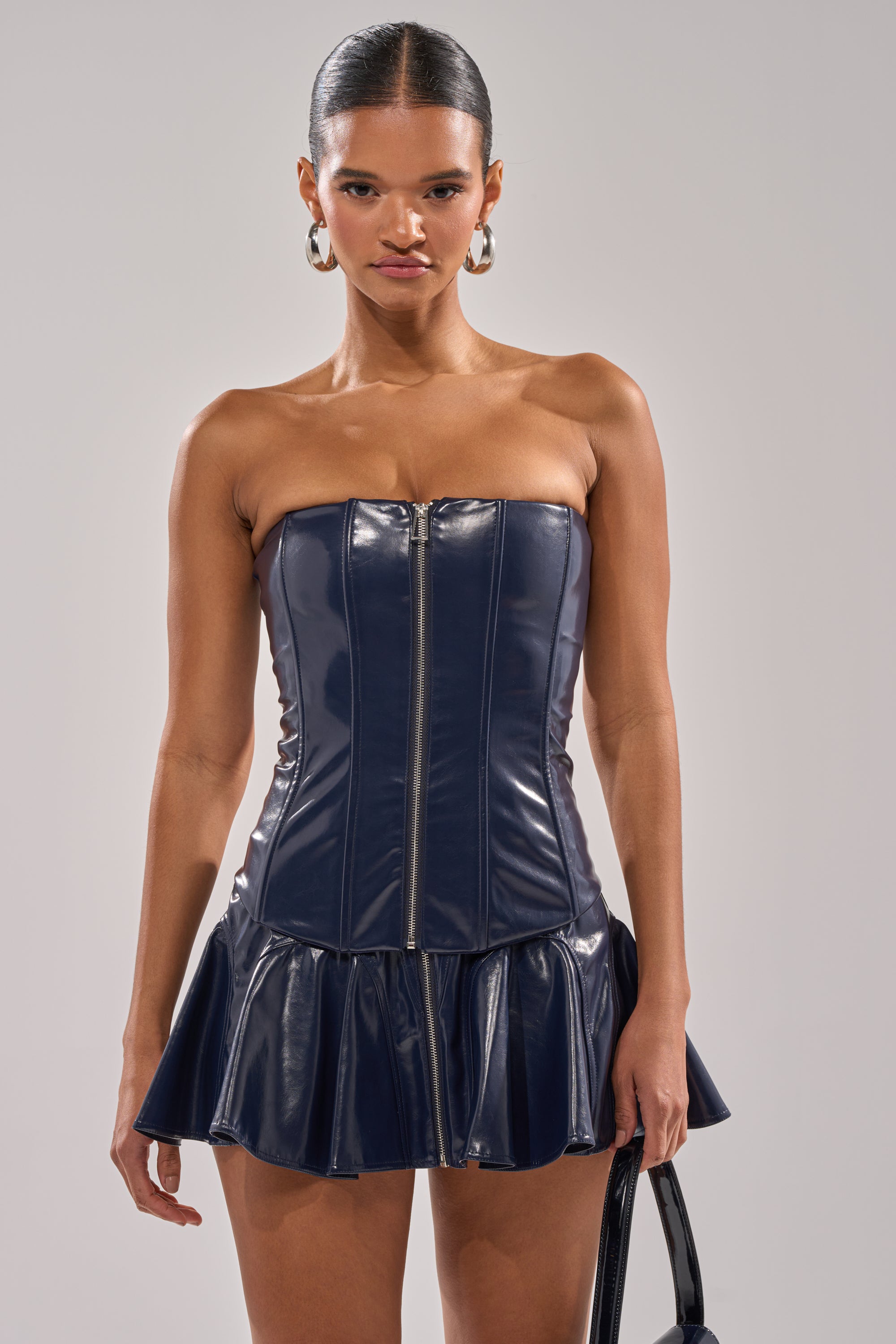A woman stands against a plain background, wearing the COME PARTY WITH ME CORSET—a navy faux patent leather strapless mini dress with a front zipper and flared skirt. She has slicked-back hair and large hoop earrings. 0