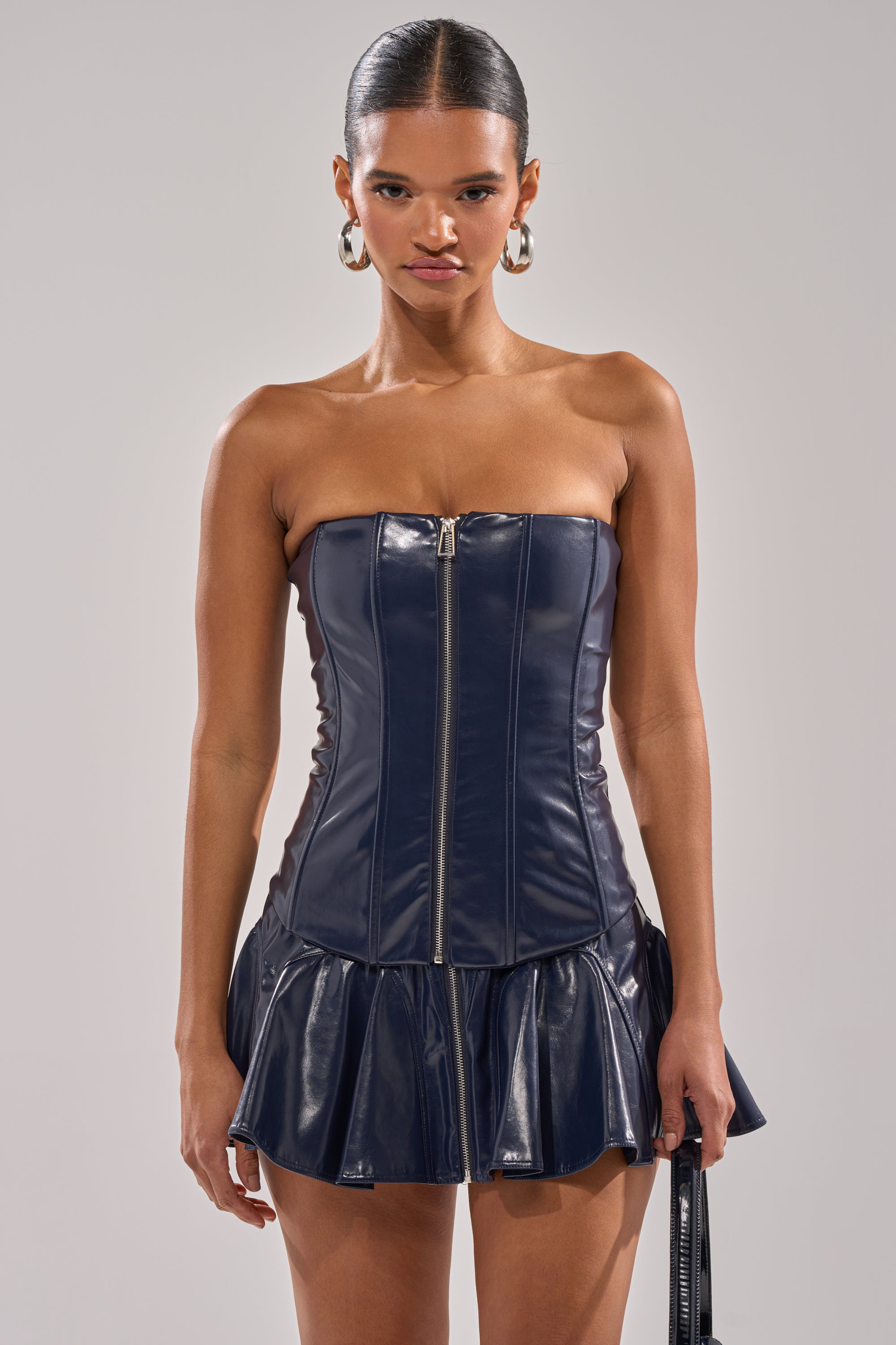 A woman models the COME PARTY WITH ME CORSET, a strapless navy blue faux leather mini dress with a front zipper and flared skirt. She stands against a plain background, her hair slicked back and hoop earrings as accessories. 2