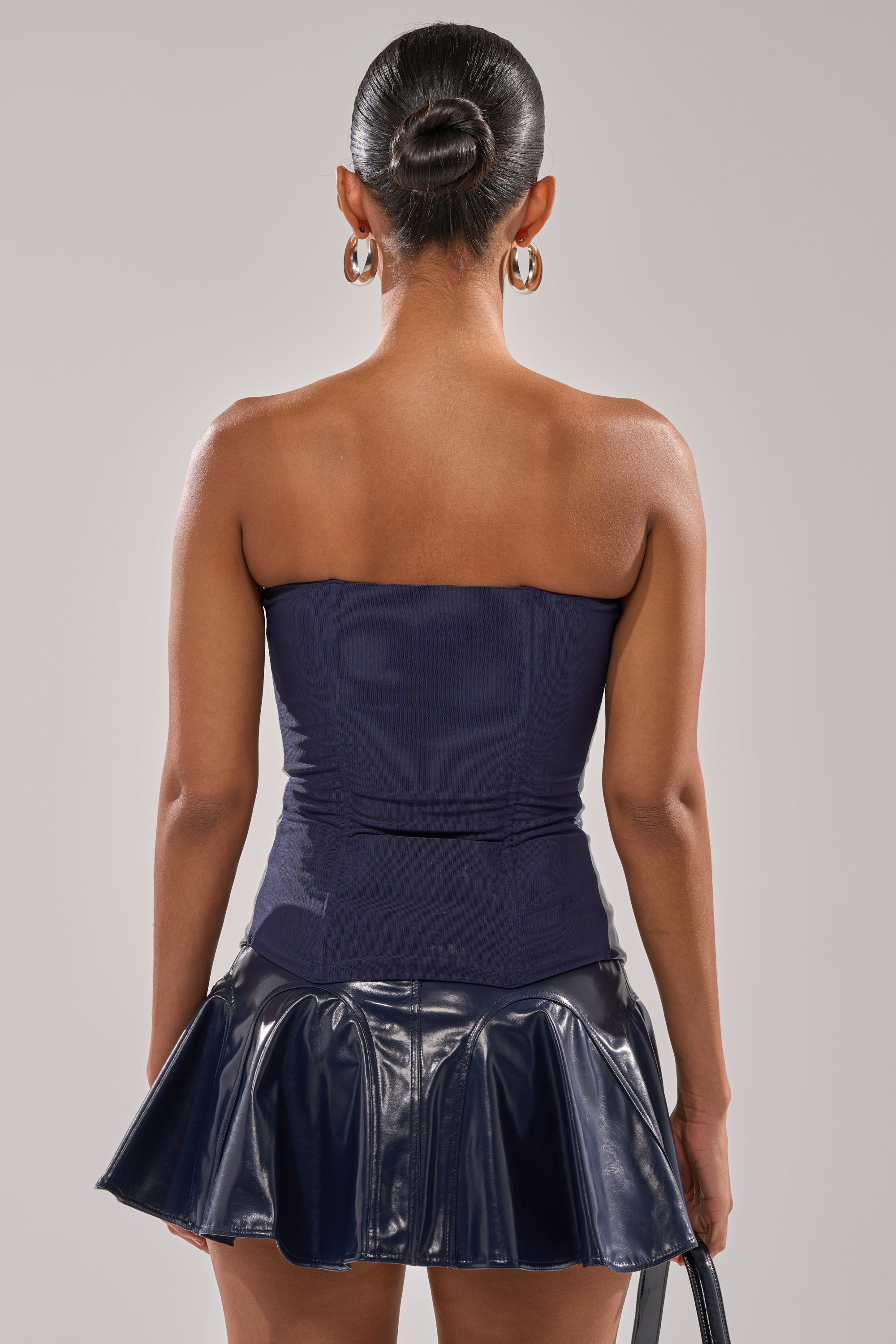 A woman faces away, revealing her upper back as she wears the COME PARTY WITH ME CORSET, a strapless navy blue top, paired with a shiny dark blue mini skirt, large hoop earrings, and her hair styled in a neat bun. 4