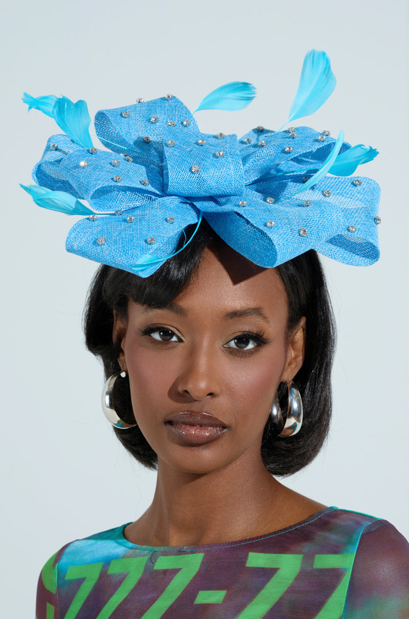 A woman wears the DENIM AND DIAMONDS CLIP ON HEADBAND, a vibrant blue fascinator with rhinestones and feather details. She has sleek dark hair and silver hoop earrings, pairing it with a multicolored abstract patterned top against a light blue background.