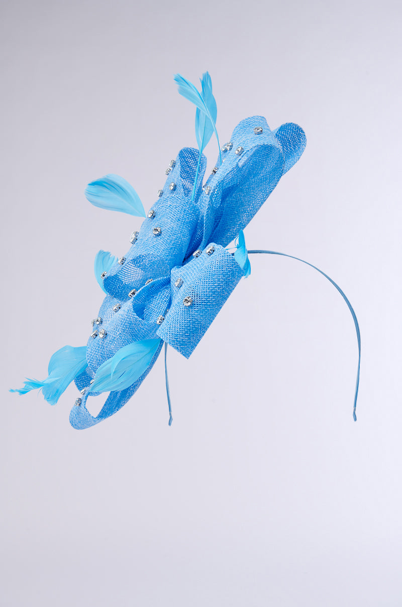 The Denim and Diamonds Clip On Headband features a blue fascinator with a mesh bow, rhinestone accents, feather details, and a thin band, perfect for adding flair to any outfit against a plain background.