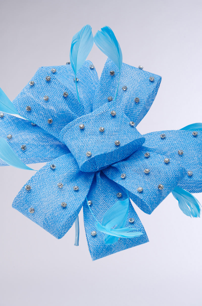 A vibrant blue fabric bow, like a Denim and Diamonds Clip-On Headband, adorned with rhinestones and delicate blue feathers, is set against a plain light background.