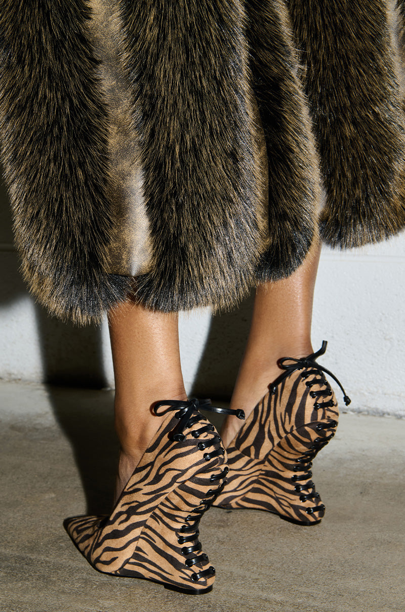 A person wearing the AZALEA WANG KABIRA MULTI WEDGE PUMP with tiger-print lace-up high heels featuring wavy designs stands on a concrete floor. The shoes showcase unique, faux suede details. Part of a furry, brown-striped coat is visible, draping near the person's ankles.