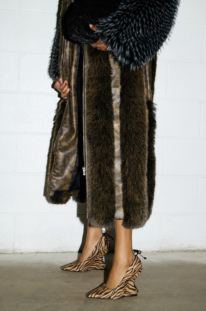 Dressed in a long faux fur coat, the person holds a black bag while wearing the AZALEA WANG KABIRA MULTI WEDGE PUMP, featuring zebra-patterned pointed-toe heels with ribbon laces. They are standing against a white brick wall, exuding an edgy yet sophisticated style that seamlessly blends urban chic and wild flair akin to tiger print accents.