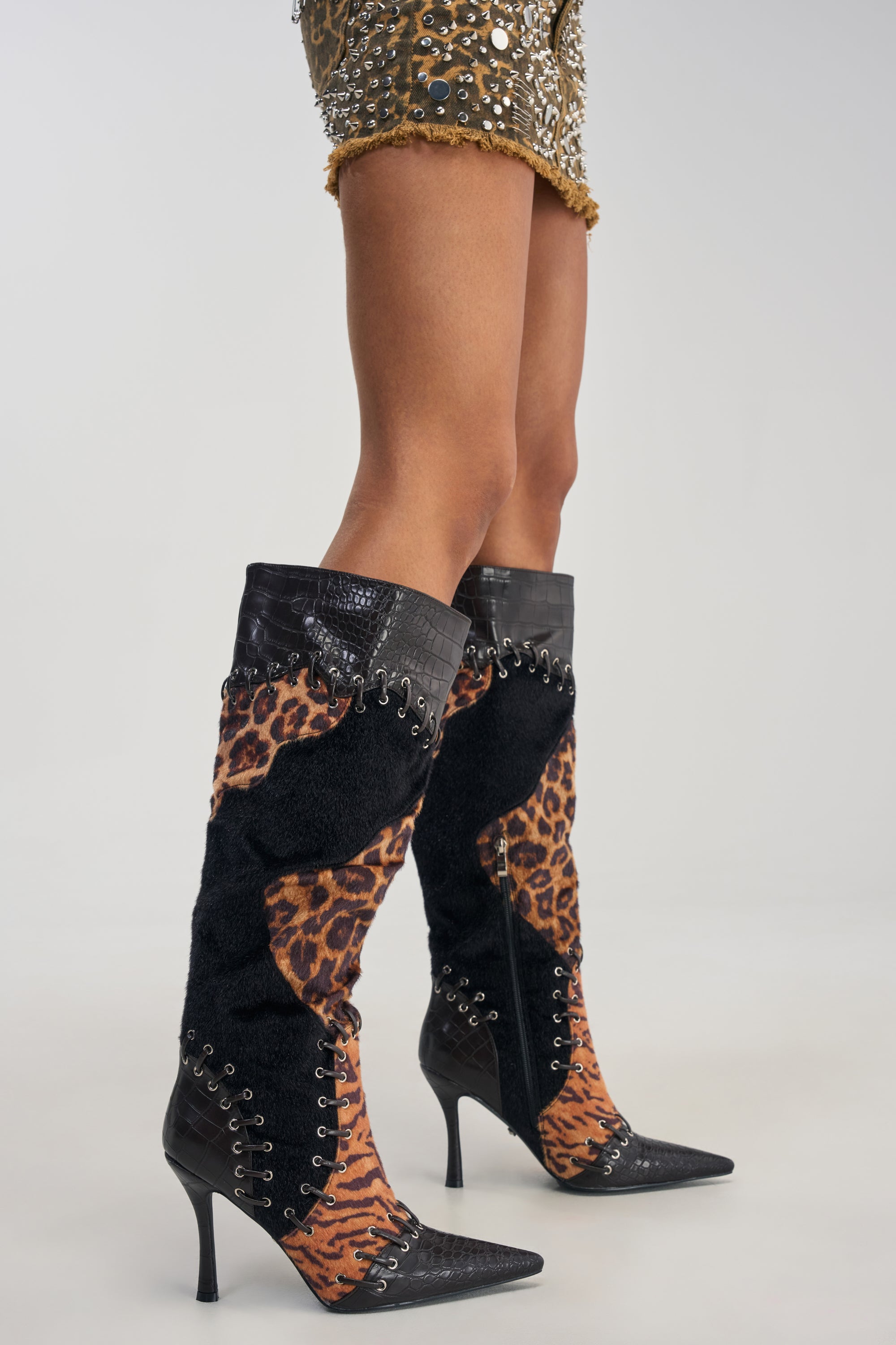A person wearing the AZALEA WANG KAIROX ANIMAL PATCHWORK LUXE BOOT—stylish knee-high stiletto boots with black laces and pointed heels in an eye-catching animal print—paired with a short studded skirt.