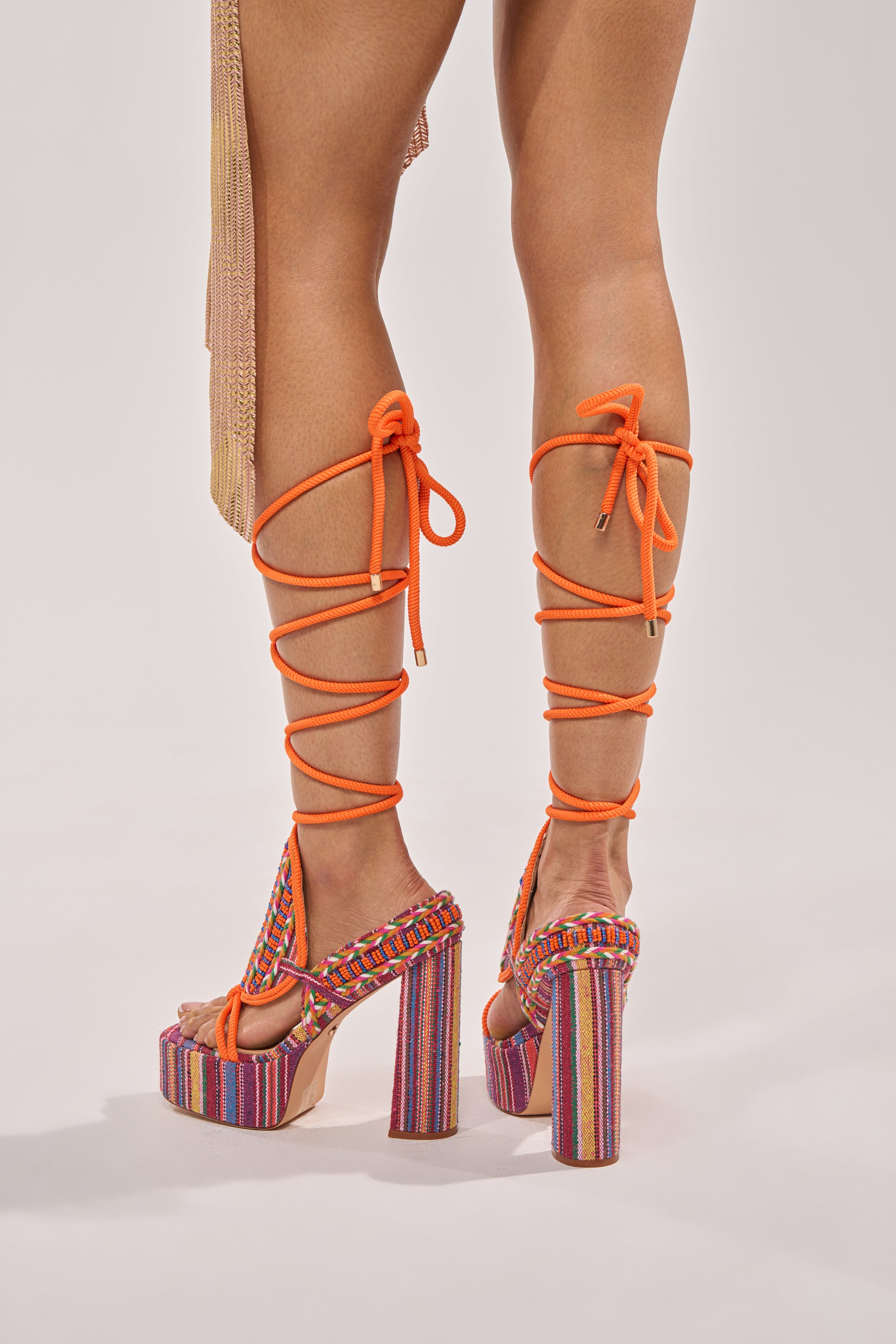 A person wears the AKIRA GOLD KALINA MULTI CHUNKY BEADED SANDAL with colorful stripes and orange lace-up straps wrapping around the calves, standing on a light background. 3