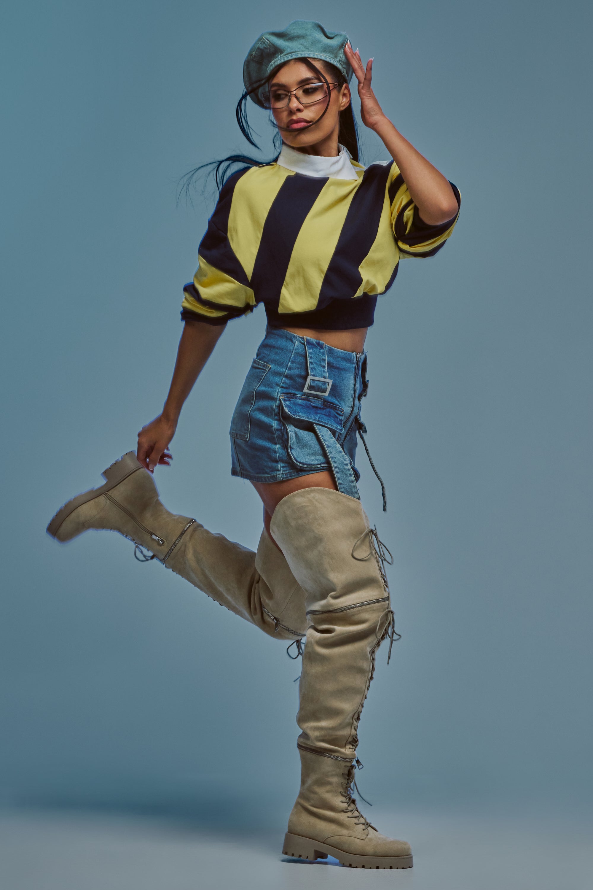 A person poses stylishly in a striped cropped sweater, denim shorts, and the AKIRA GOLD KALLIN BEIGE CONVERTIBLE SHAFT HEIGHT BOOT, accessorized with glasses and a blue beret, set against a plain blue background.