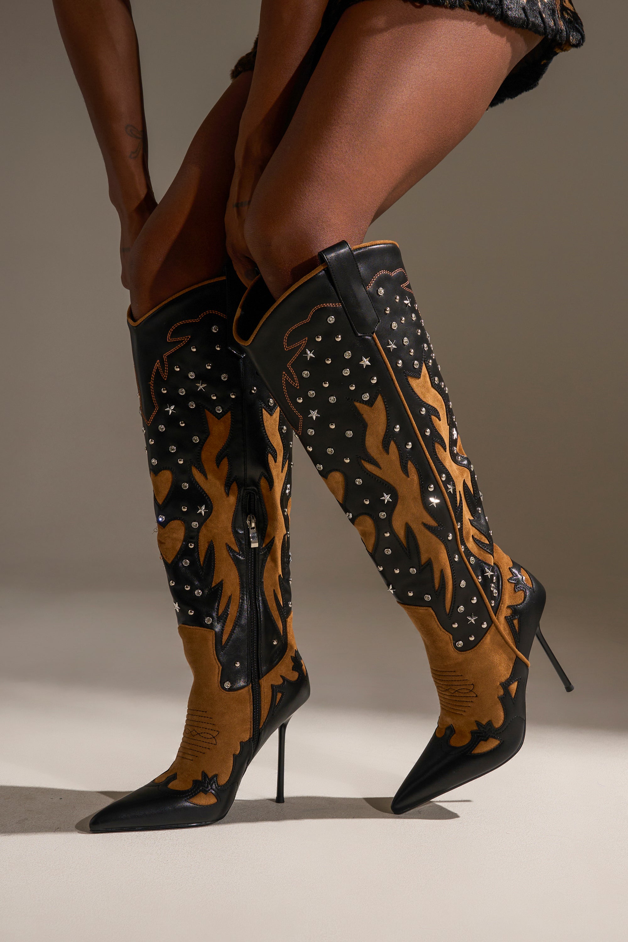 Wearing the AKIRA GOLD KALPANAS BLACK MULTI STILETTO WESTERN BOOT, featuring bold black and brown flame designs with silver studs, this person stands confidently—legs crossed, hands on thighs—showcasing true cowboy flair.