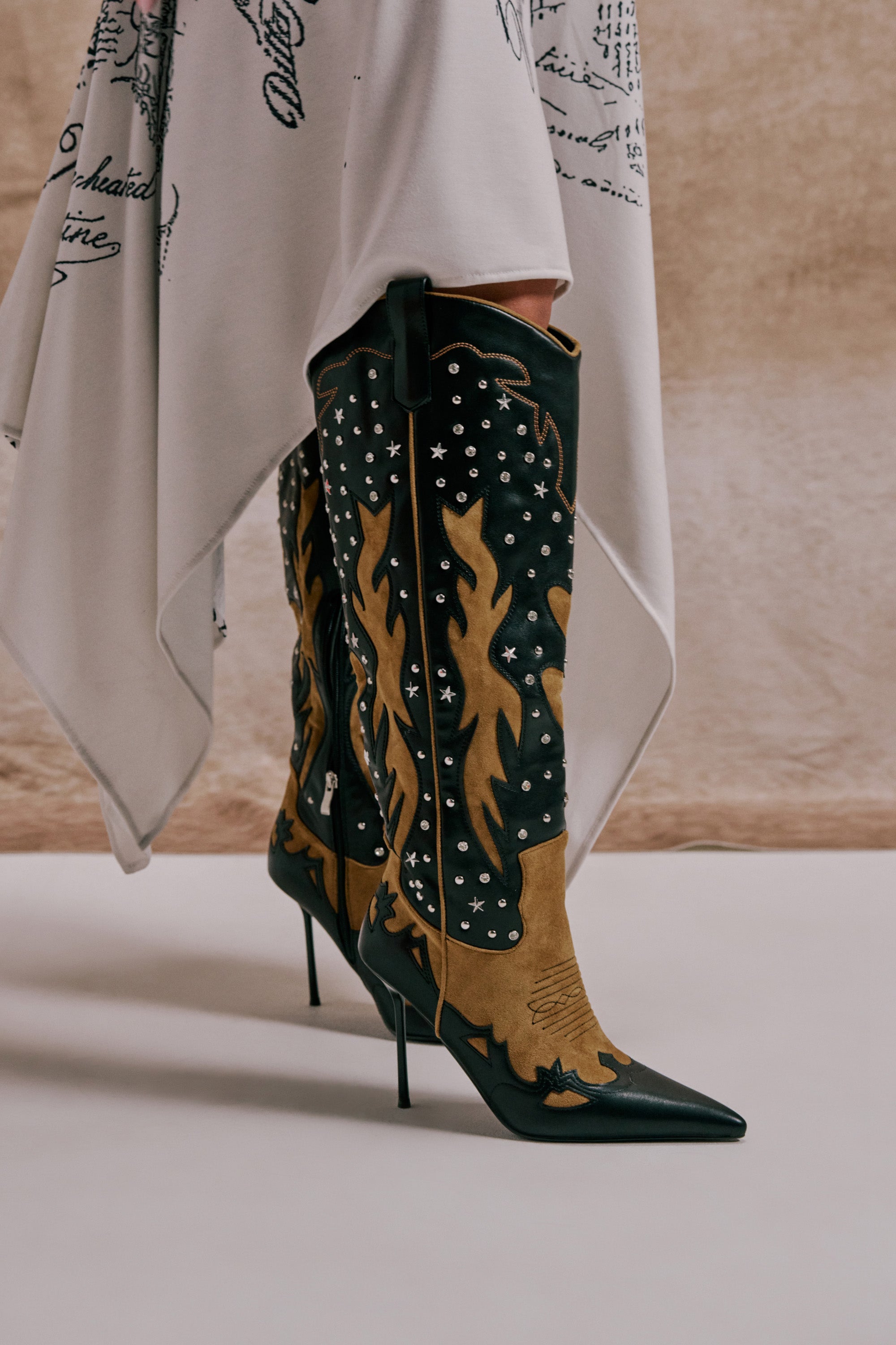 A person wears a white asymmetrical skirt and AKIRA GOLD KALPANAS BLACK MULTI STILETTO WESTERN BOOTS, featuring black-and-tan hues, flame patterns, silver studs, and pointed toes, standing on a light surface against a beige background. 0