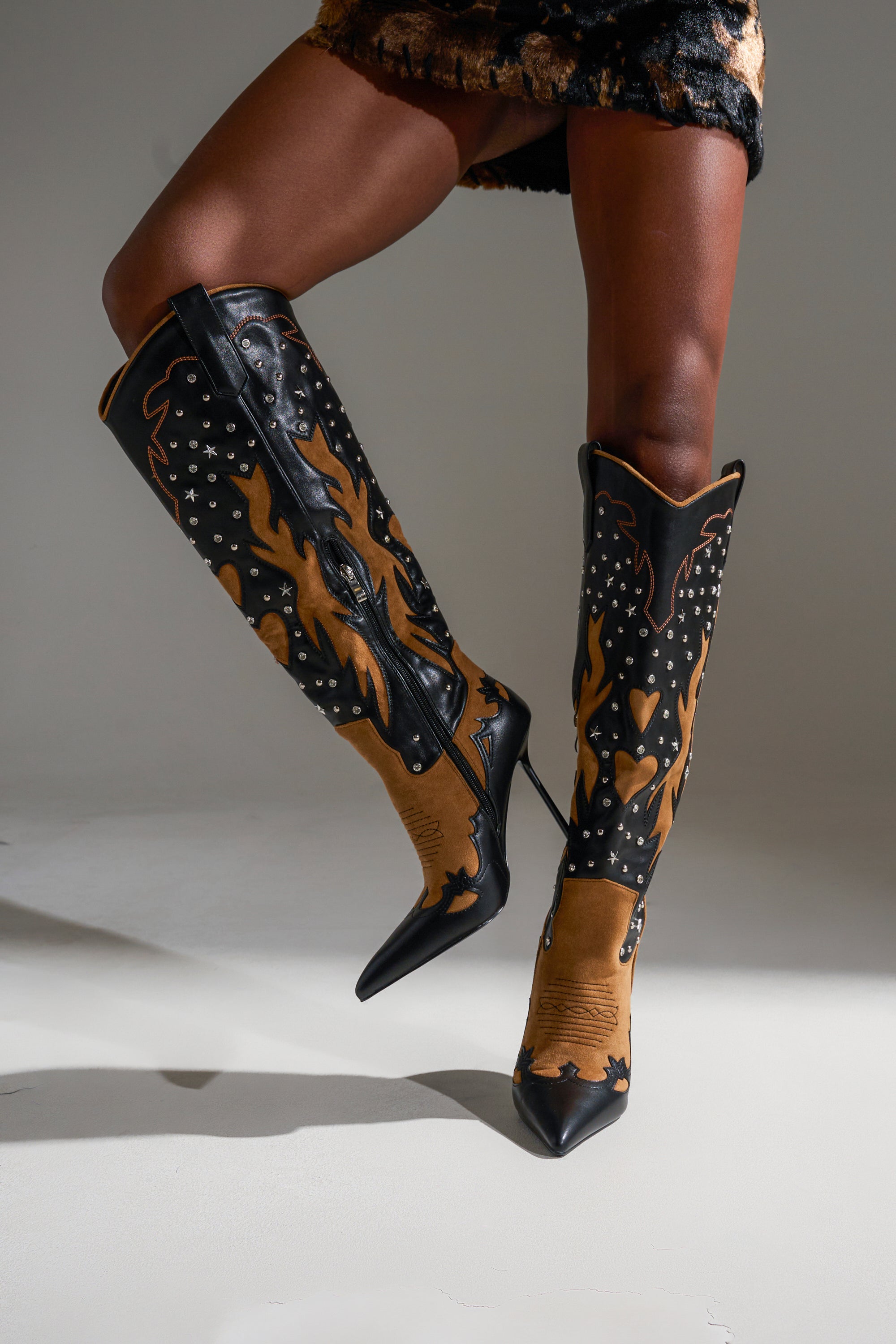 A person shows off bold cowboy flair in the AKIRA GOLD KALPANAS BLACK MULTI STILETO WESTERN BOOT, featuring knee-high, pointed toes, black and brown flame patterns, silver studs, and high heels with one leg slightly crossed.