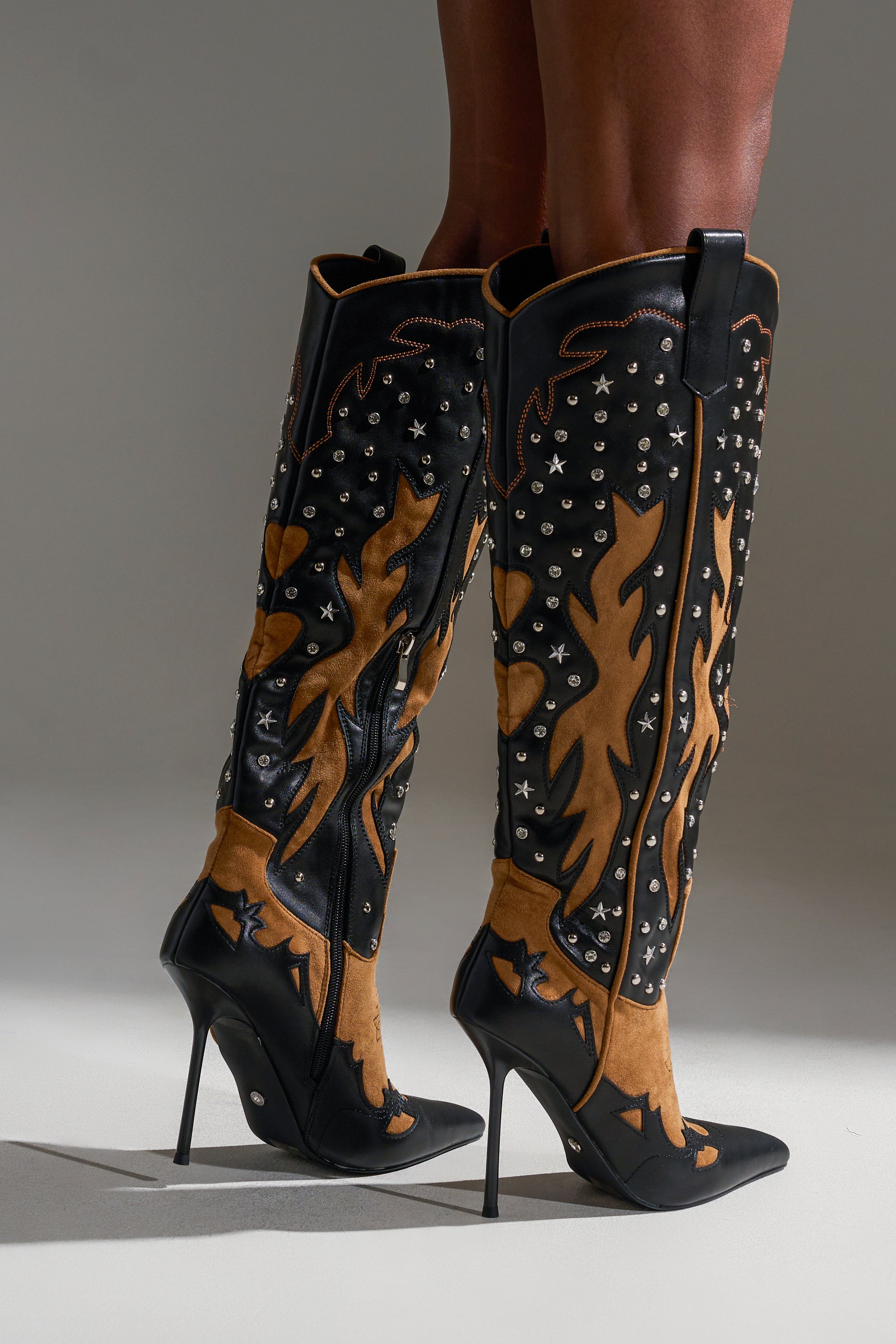 The AKIRA GOLD KALPANAS BLACK MULTI STILETTO WESTERN BOOT features black and tan leather, decorative stitching, silver studs, and flame-inspired patterns, adding true cowboy flair as worn by a person on a light background.