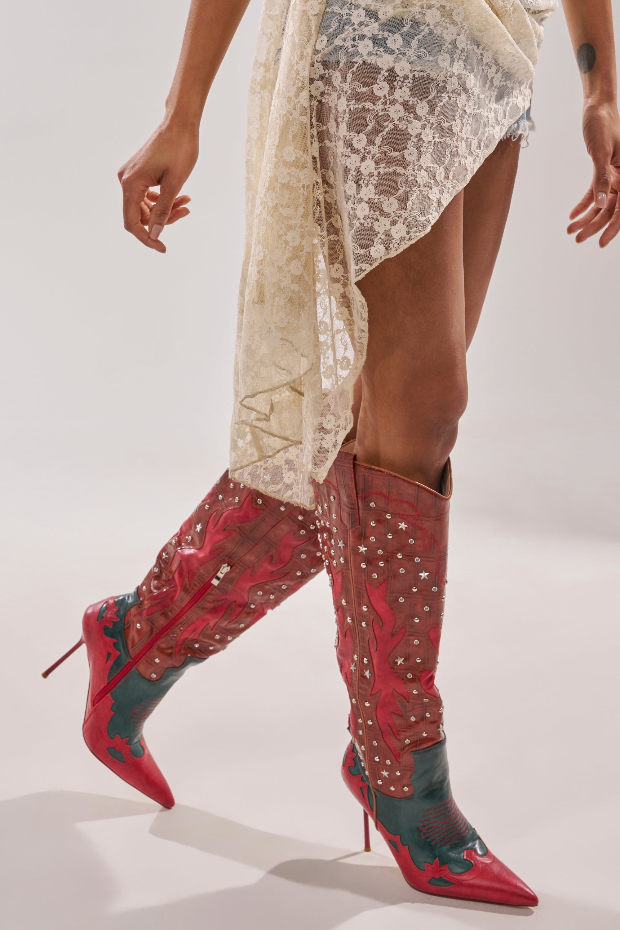 A person in a sheer off-white lace skirt walks against a light background, wearing AZALEA WANG KALPANAS MULTI WESTERN STILETTO BOOTS featuring striking red color, knee-high heels, and star-shaped studs. 7