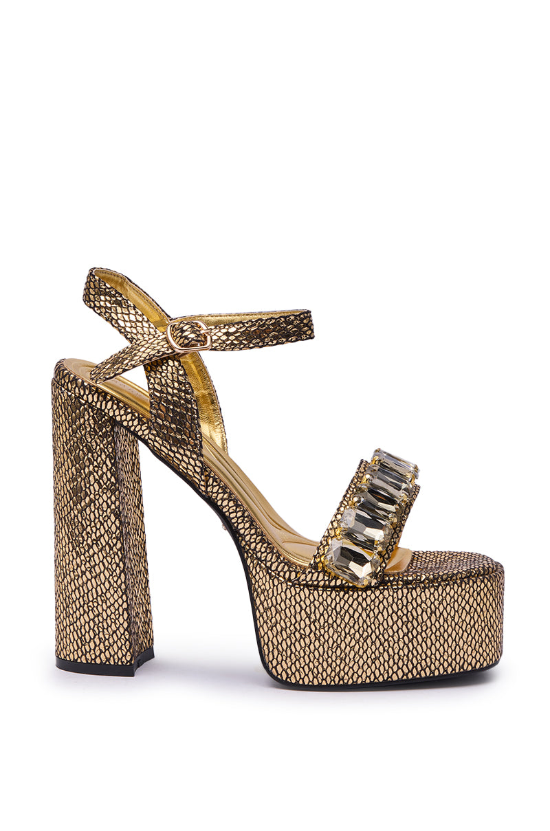 The AZALEA WANG KAMILLA GOLD RHINESTONE SANDAL has a chunky heel, platform sole, and textured surface. Large rectangular gemstones adorn the toe strap, and an ankle strap provides support and style.
