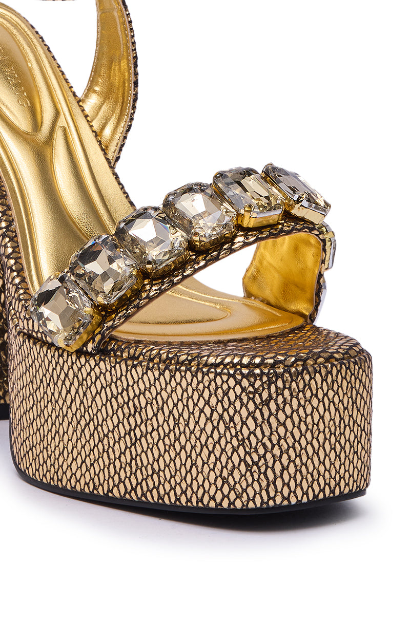 The AZALEA WANG KAMILLA GOLD RHINESTONE SANDAL features a chunky heel, thick textured platform sole, and large faceted gemstones on the front strap, offering a luxurious and glamorous look.