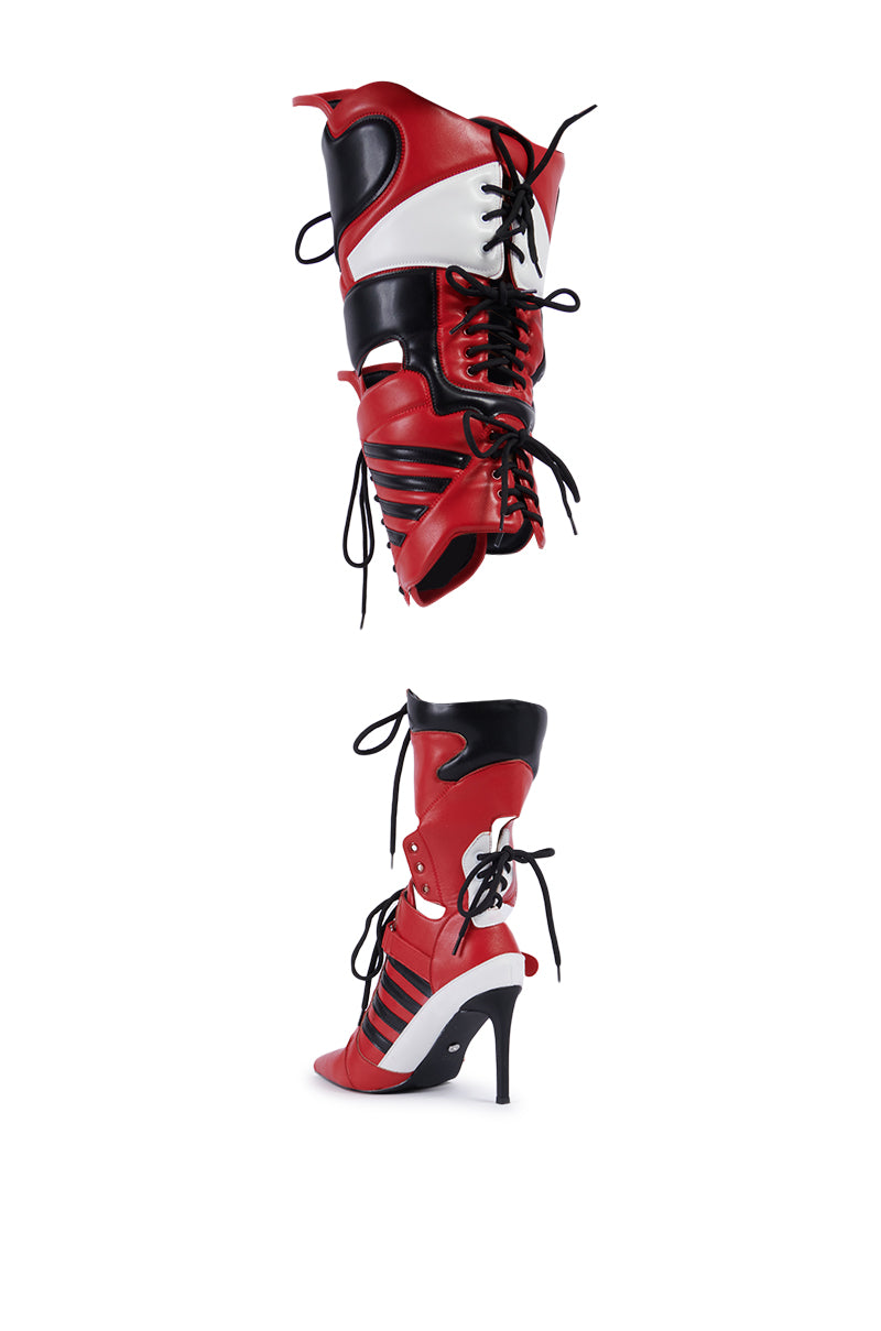 The AZALEA WANG KANEY RED SPORT 2-1 BOOT is a pair of high-heel boots with a unique design. These boots are predominantly red with black and white accents, featuring intricate laces and straps. The heels are stiletto-style, and there are cut-out details that add to the elaborate aesthetic.