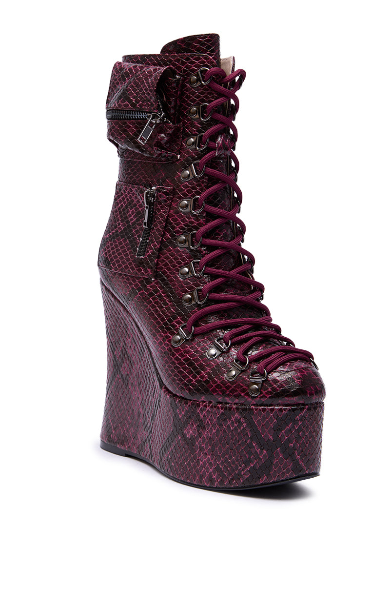 The AZALEA WANG KAPULE BURGUNDY SNAKE PLATFORM BOOTIE is a striking mid-calf, lace-up boot featuring a maroon and black snakeskin design, metal eyelets, side zipper, and a chic ankle strap with buckle on a high wedge heel.