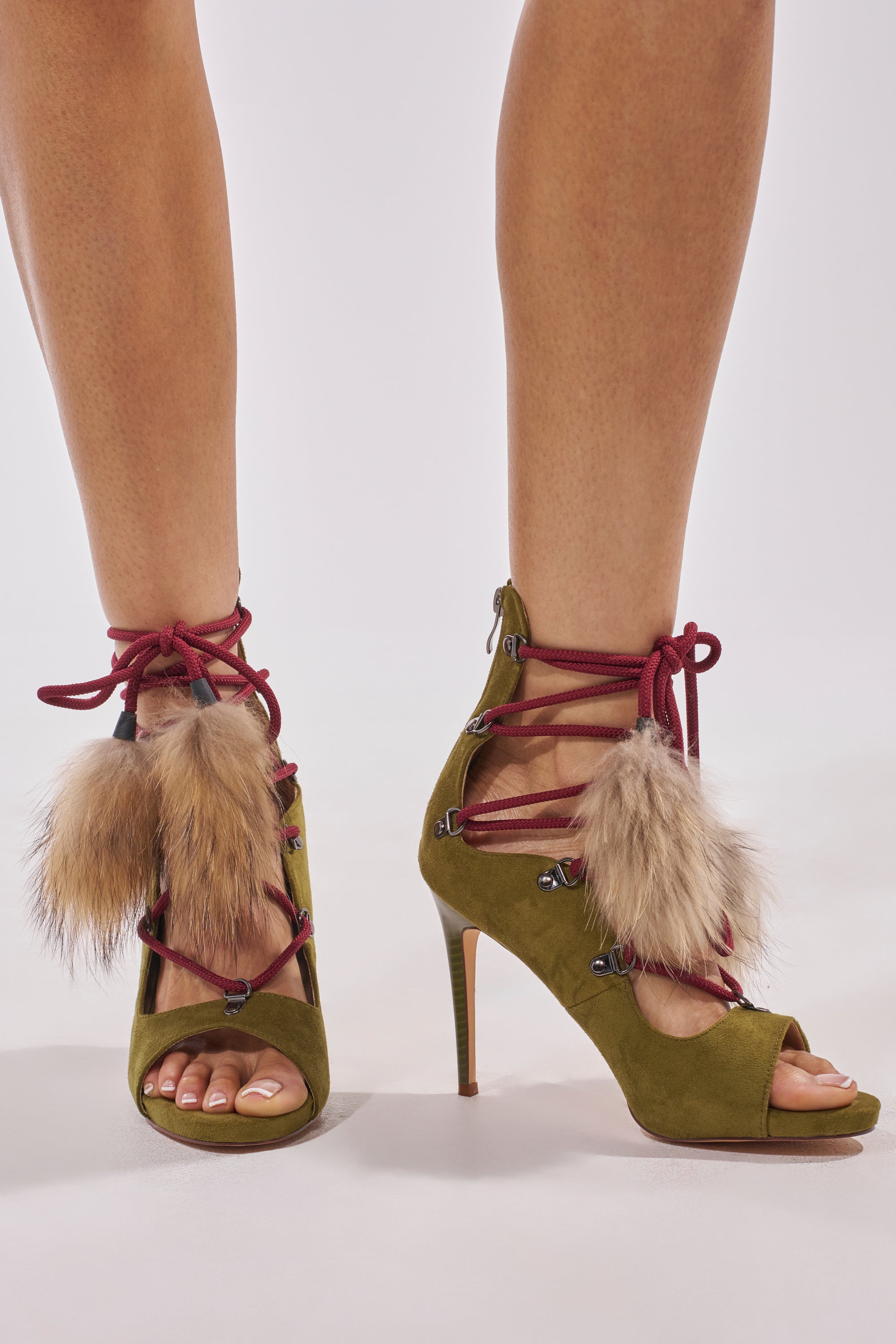 A close-up of someone wearing the AZALEA WANG KARLA OLIVE FAUX SUEDE FUR EMBELLISHED STILETTO SANDAL, featuring red laces and faux fur pompoms on the front, against a plain light background. 2