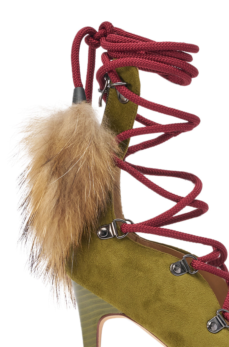 A close-up of the AZALEA WANG KARLA Olive Faux Suede Fur Embellished Stiletto Sandal showcases green suede, red laces with metal eyelets, and a brown fur pom-pom detail near the ankle. 7