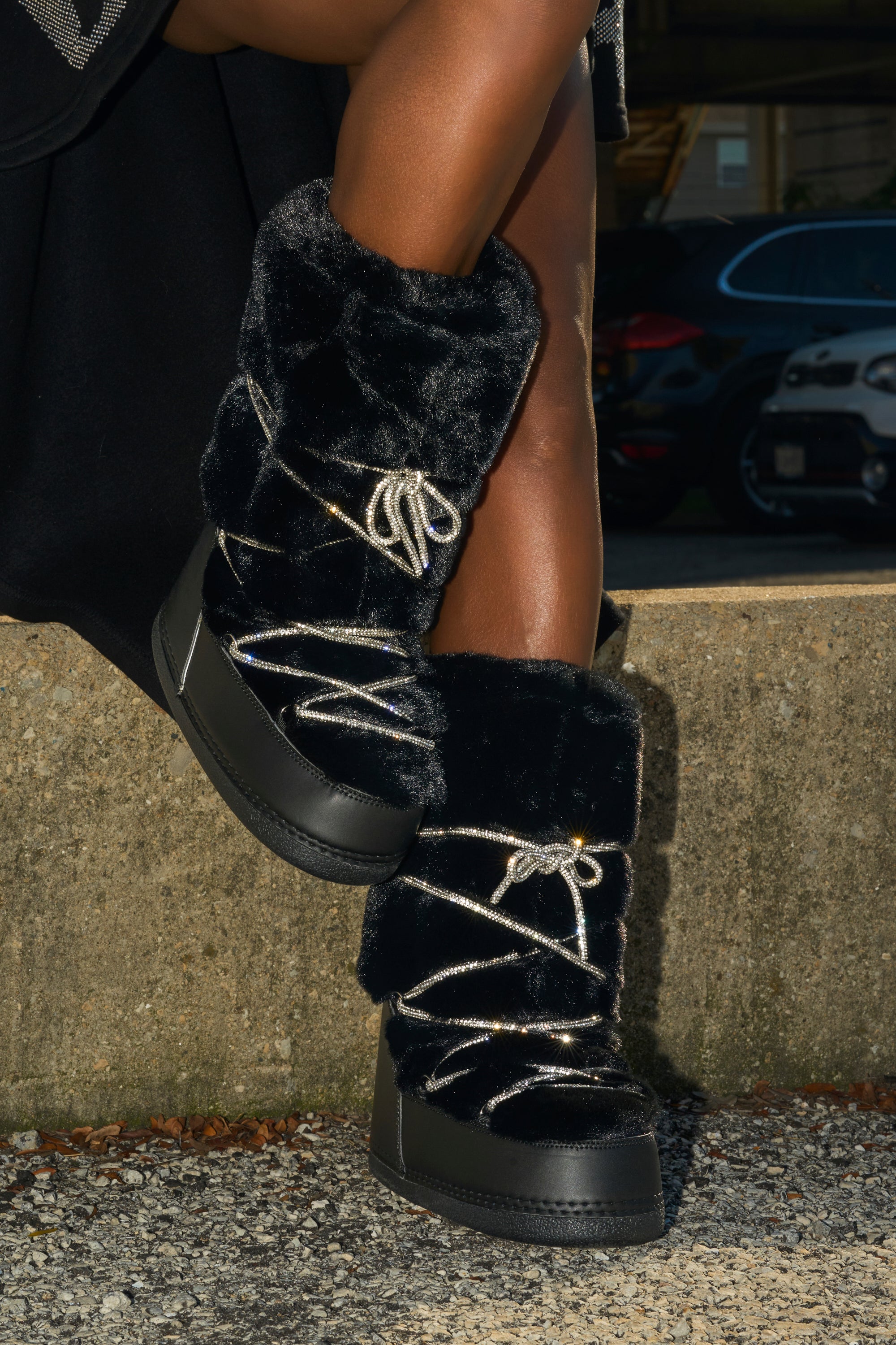 A person wearing AZALEA WANG KARMELA BLACK FUR MOON BOOTS with silver laces and thick faux fur sits cross-legged on a concrete ledge outside, with parked cars visible in the background.