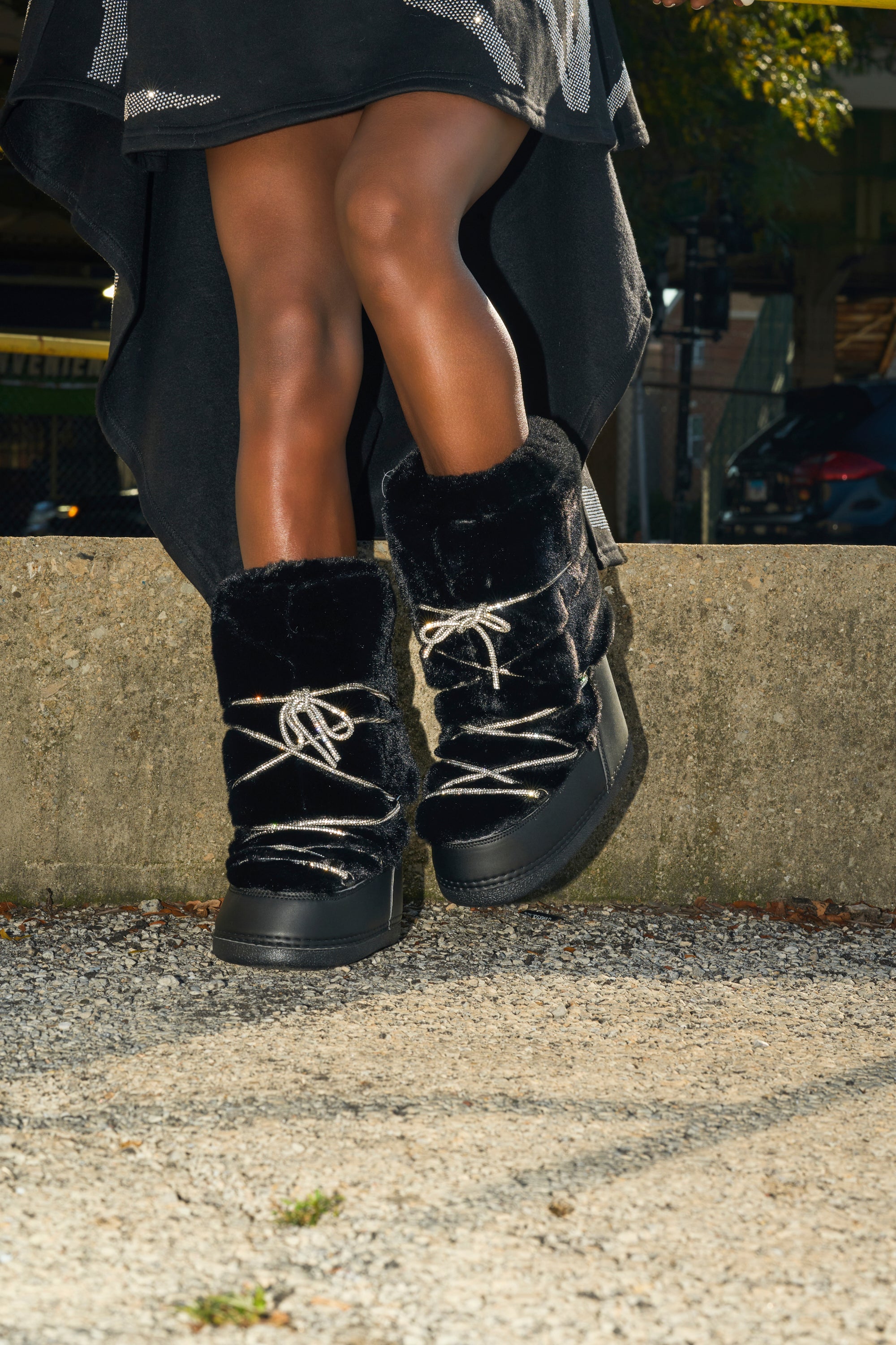 A person with dark skin stands on a gravel path by a concrete wall, wearing a black skirt and cozy AZALEA WANG KARMELA BLACK FUR MOON BOOTS with white laces. Only their legs and feet in the boots are visible.