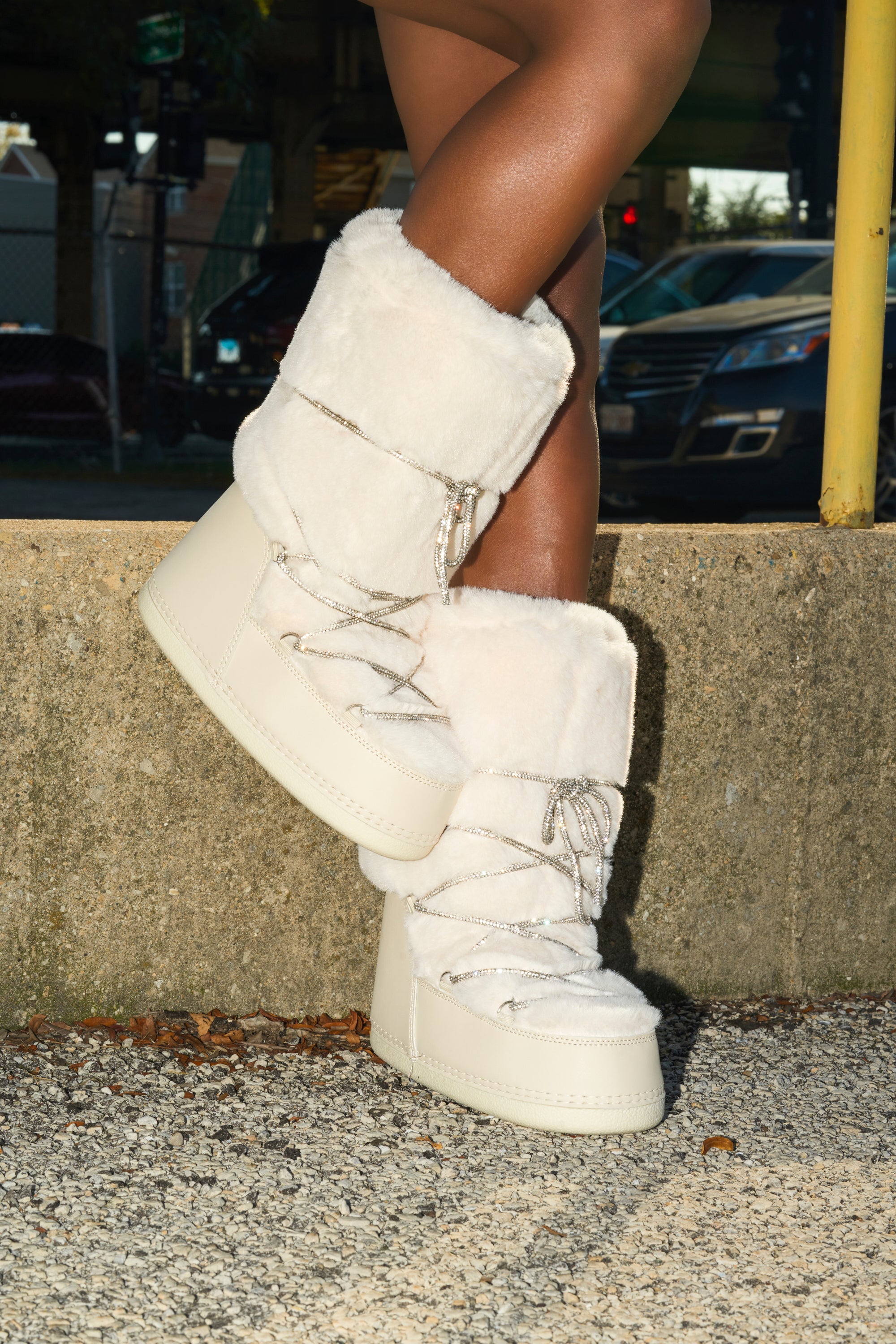 Leaning against a concrete ledge, a person stands outdoors with legs crossed at the ankle, wearing AZALEA WANG KARMELA CREAM FUR MOON BOOTS—tall, cream boots with thick soles and lace-up detail.