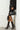A person leans against a white brick wall wearing a studded black jacket, black bicycle shorts with a bow detail, sheer socks, and the AZALEA WANG KASHIE BLACK STILETTO PUMP. They hold a black handbag adorned with metal spikes and eyelets.