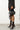 A person stands against a white brick wall, wearing a studded black jacket, black shorts with ribbon details, and the AZALEA WANG KASHIE BLACK STILETTO PUMP with pointed toes. They hold a small black handbag adorned with metal eyelets.
