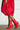 A person stands against a white brick wall, adorned in a bright red dress with voluminous textures and sleek faux leather boots. The footwear, known as the AZALEA WANG KATHERINE RED PU BOOTIE, showcases side zippers and complements the minimalist concrete floor of the indoor setting.