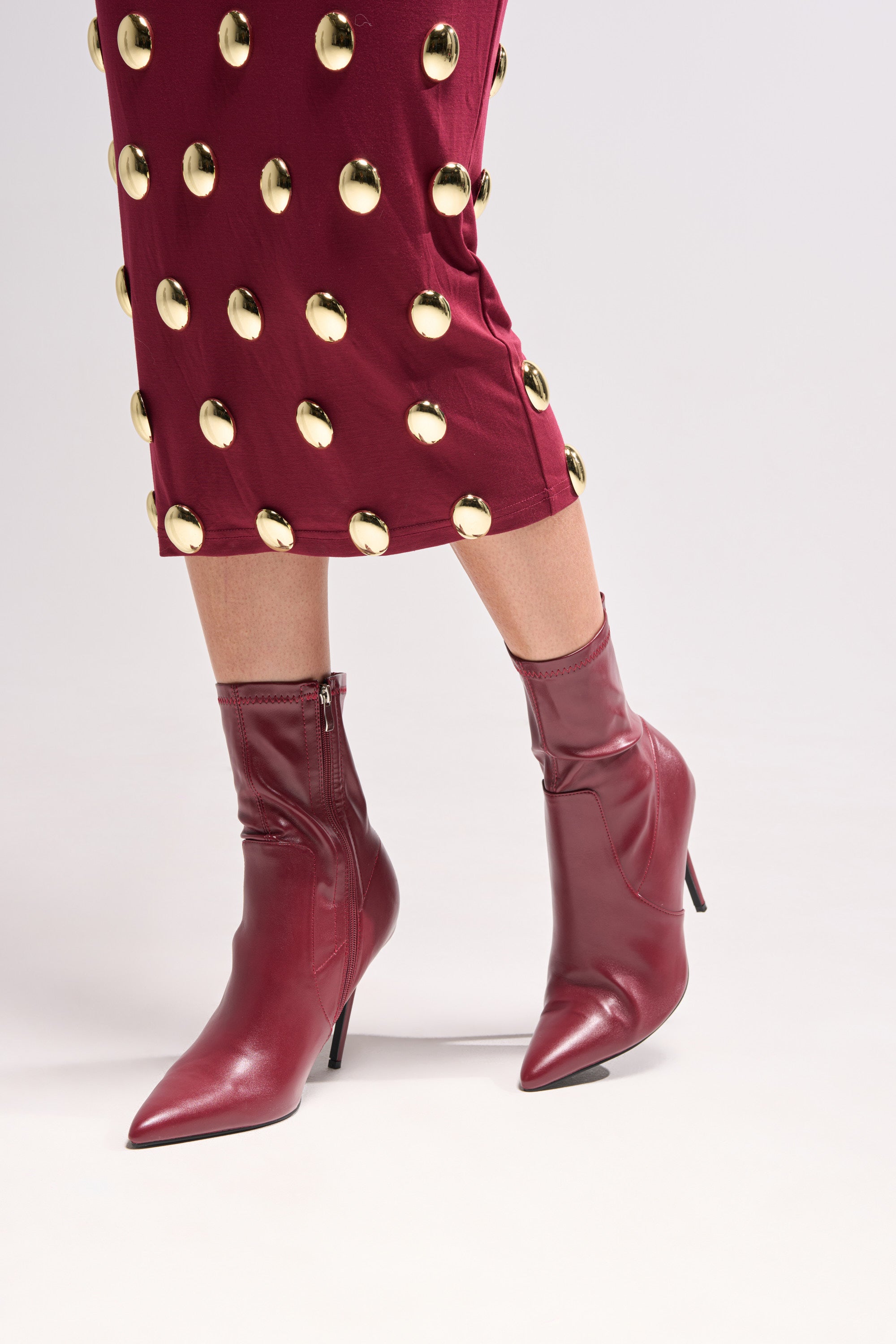 Someone stands on a light surface wearing a maroon skirt with large gold circular studs and the AZALEA WANG KATHII BURGUNDY BOOTIE, which features a stiletto heel for a matching, stylish look.