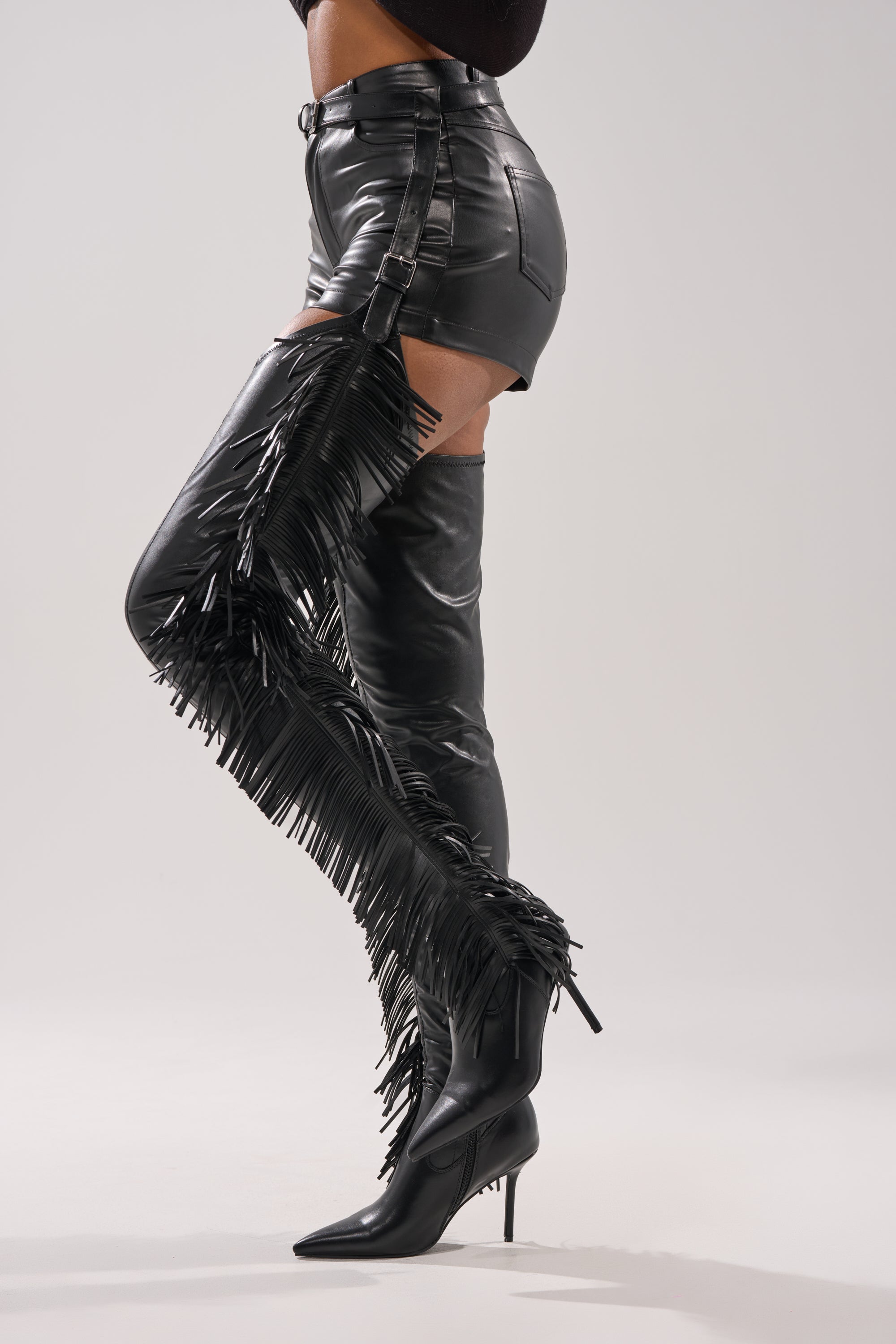A person wears black faux leather shorts and AZALEA WANG KATRICE Black Stiletto Western Fringe Chap Boots, standing against a plain light background. 3