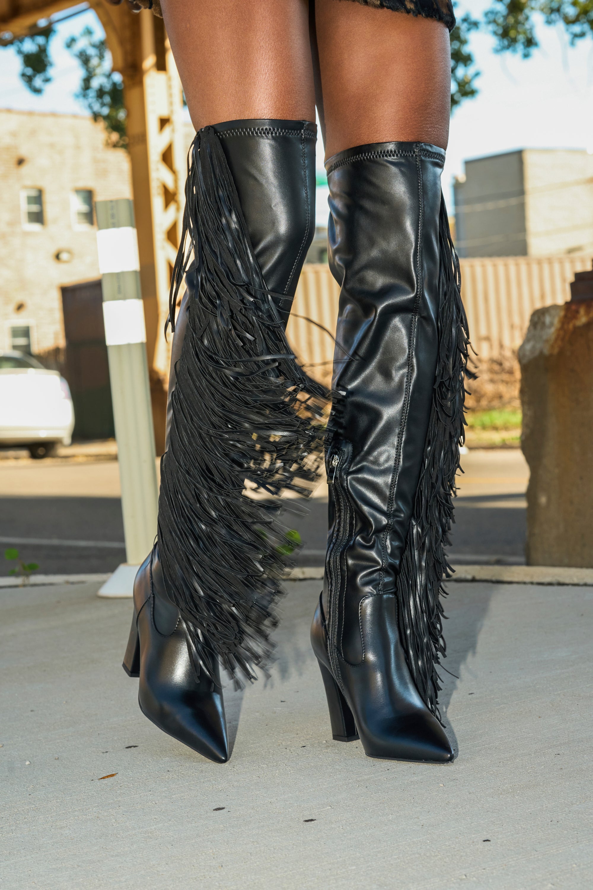 A person stands outdoors on a sidewalk wearing the AZALEA WANG KATRICIA BLACK FRINGE CHUNKY BOOT; urban buildings appear in the background.