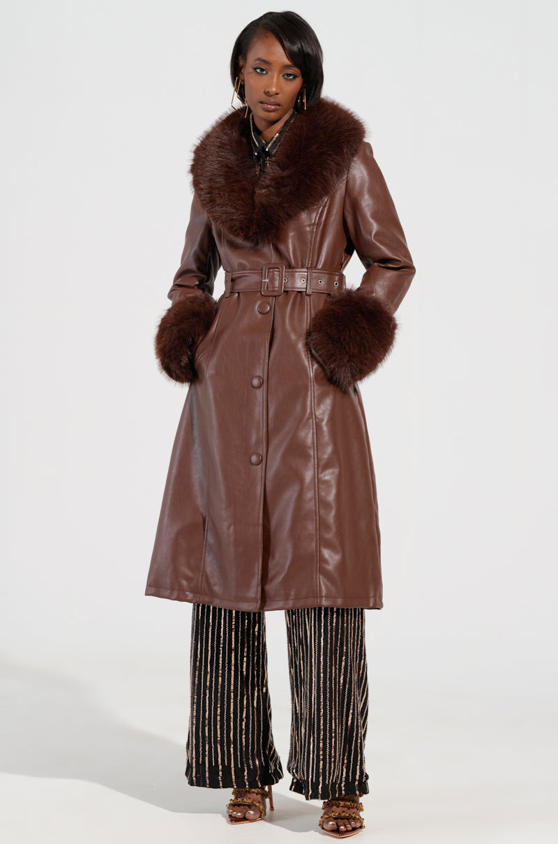 A person is dressed in the Kaya Fur Lined Trench in brown, featuring a faux leather design with a faux fur collar and cuffs, belted at the waist. They complement it with patterned pants and open-toed heels on a plain, light background.