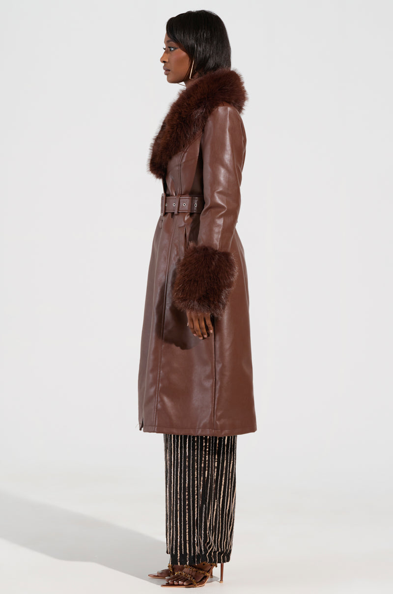 A person is modeling the KAYA FUR LINED TRENCH IN BROWN, which features a long brown faux leather design with a sherpa lining and fur collar. They are styled in patterned pants and pointy heels against a solid white backdrop.