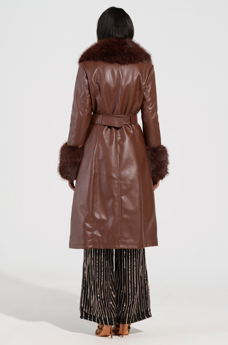 A rear view of a person wearing the KAYA FUR LINED TRENCH IN BROWN, featuring a long faux leather design with a sherpa lining and an elegant fur collar. The belted coat enhances their patterned skirt and brown shoes, set against a plain background and radiating timeless elegance.