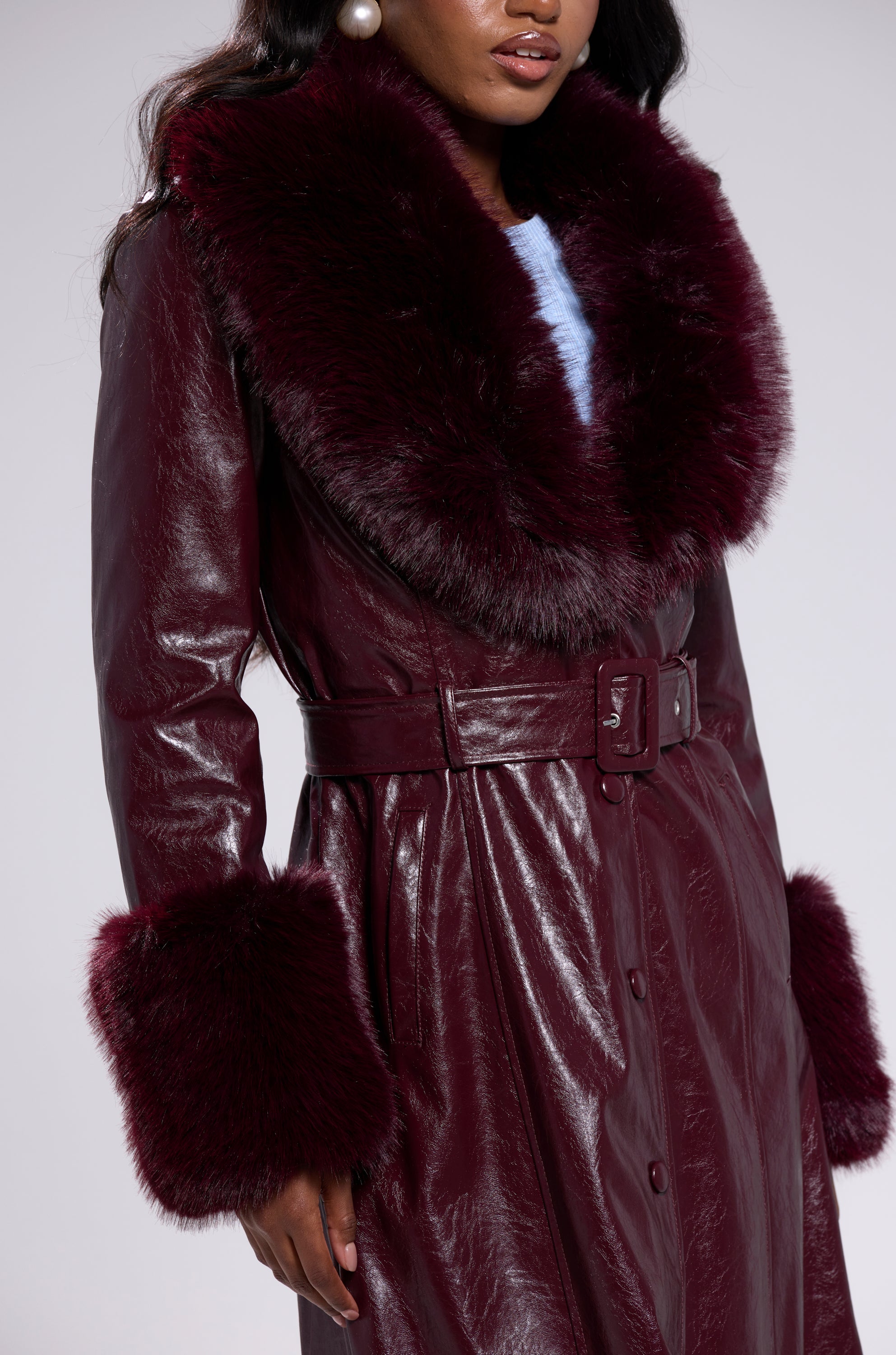 A woman wears the KAYA FUR TRIM TRENCH IN BURGUNDY, a glossy vegan leather coat with a wide faux fur collar and cuffs, belted at the waist. She has pearl earrings and her face is partly visible.