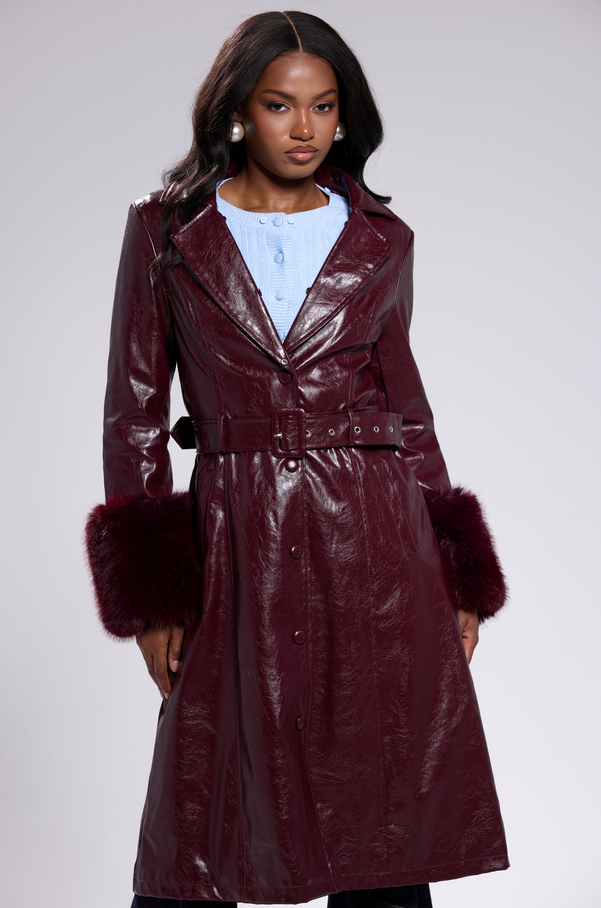 A woman wears the KAYA FUR TRIM TRENCH IN BURGUNDY, featuring a shiny finish, belted waist, and dark faux fur trim cuffs, layered over a light blue top against a plain, light background.