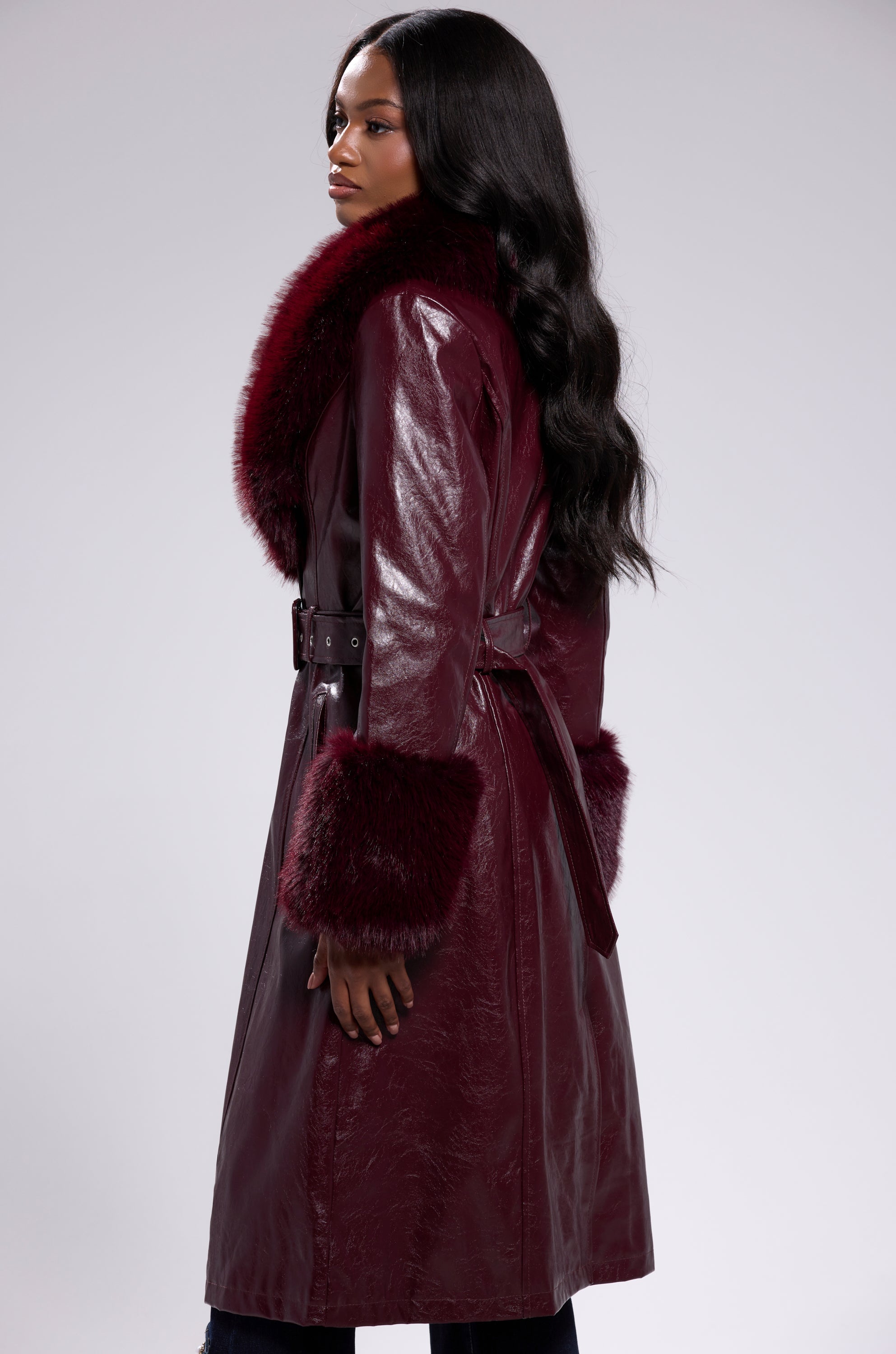 A woman with long, wavy dark hair models the KAYA FUR TRIM TRENCH IN BURGUNDY, featuring shiny fabric, a wide faux fur collar, and matching cuffs, shown in profile against a plain light background.
