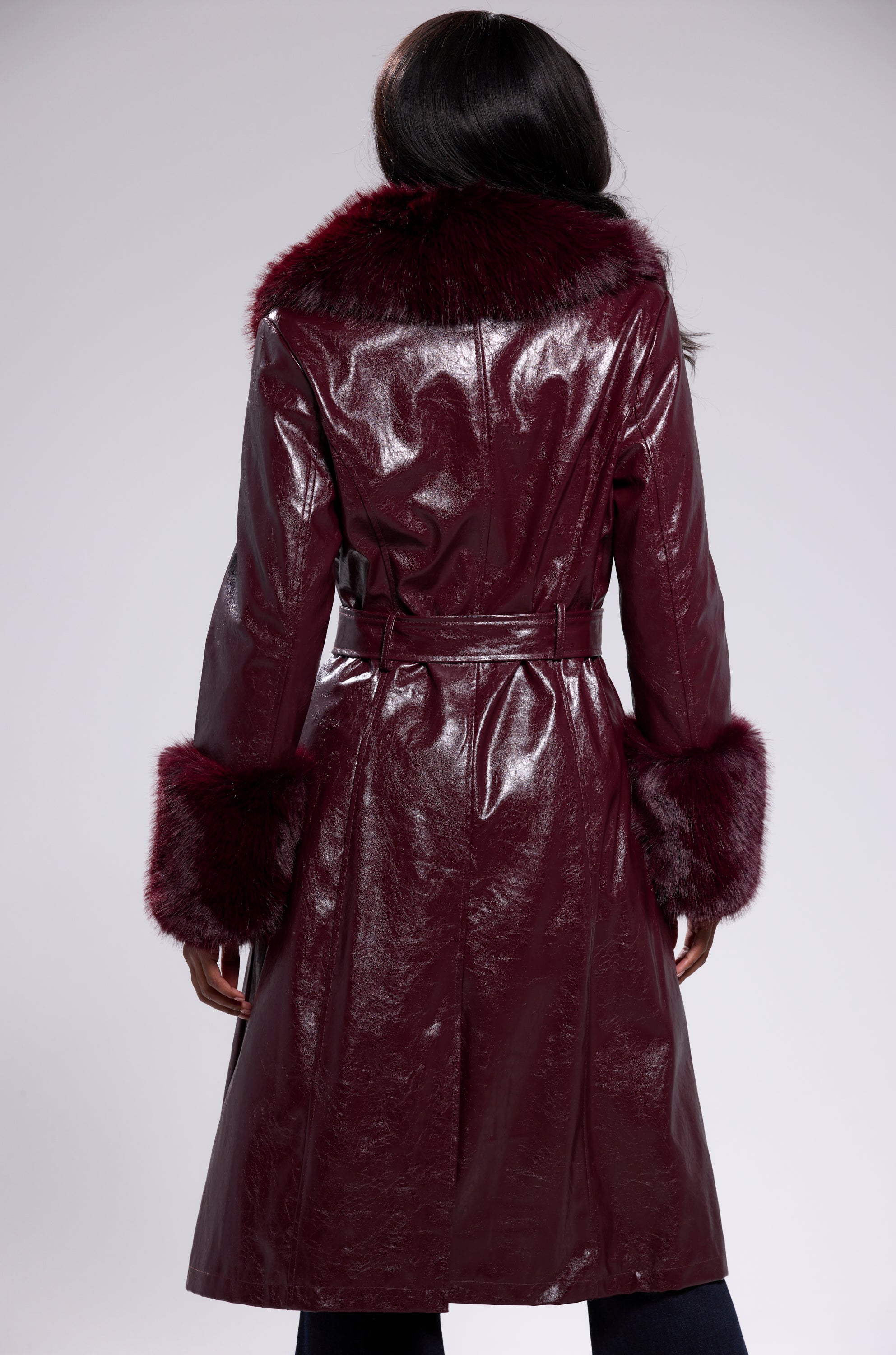 A person with long dark hair models the KAYA FUR TRIM TRENCH IN BURGUNDY, featuring faux fur collar and cuffs, viewed from the back against a plain gray background.