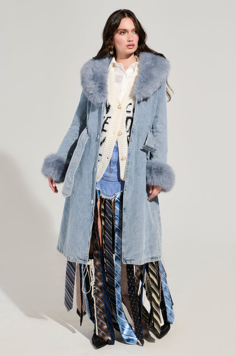 Wearing the KAYA FUR TRIM TRENCH IN DENIM featuring a sherpa lining and faux fur collar over a white patterned sweater, a person stands against a plain backdrop, highlighted by their unique skirt crafted from assorted neckties.