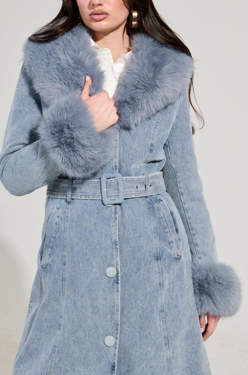 A woman lightly holds the faux fur collar of the KAYA FUR TRIM TRENCH IN DENIM. The trench has a matching belt, large buttons, and perfectly complements her long dark hair as she stands against a plain, light-colored background.