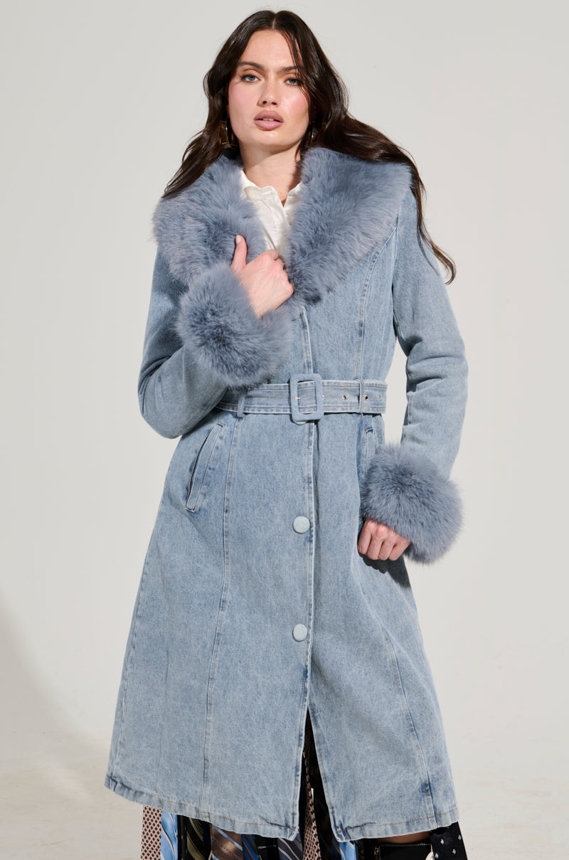 Someone wearing the KAYA FUR TRIM TRENCH IN DENIM, featuring a light blue denim finish with a faux fur collar and cuffs, poses against a plain background. The coat includes belt and button closures, and they rest one hand near the collar.