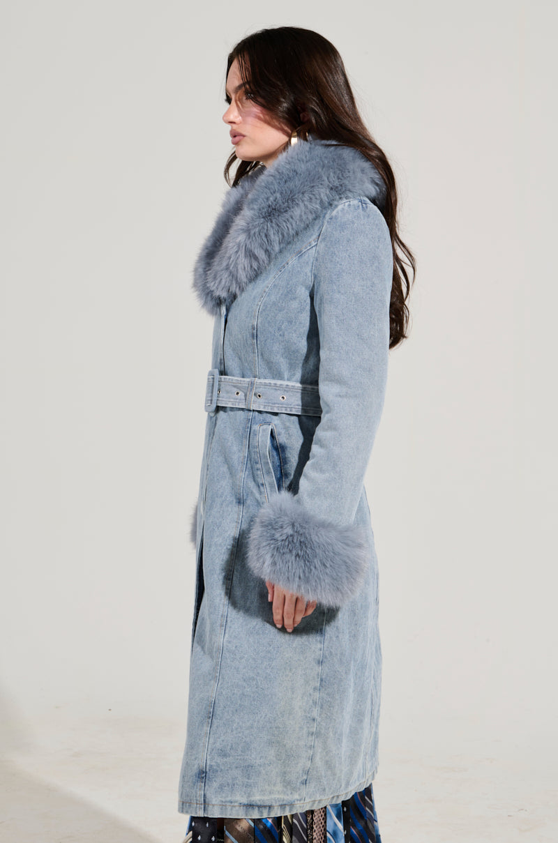 Someone models the KAYA FUR TRIM TRENCH IN DENIM against a plain backdrop, showcasing its long silhouette with a fluffy faux fur collar and cuffs. The belted design includes blue-gray fur accents and sherpa lining, adding warmth and style.