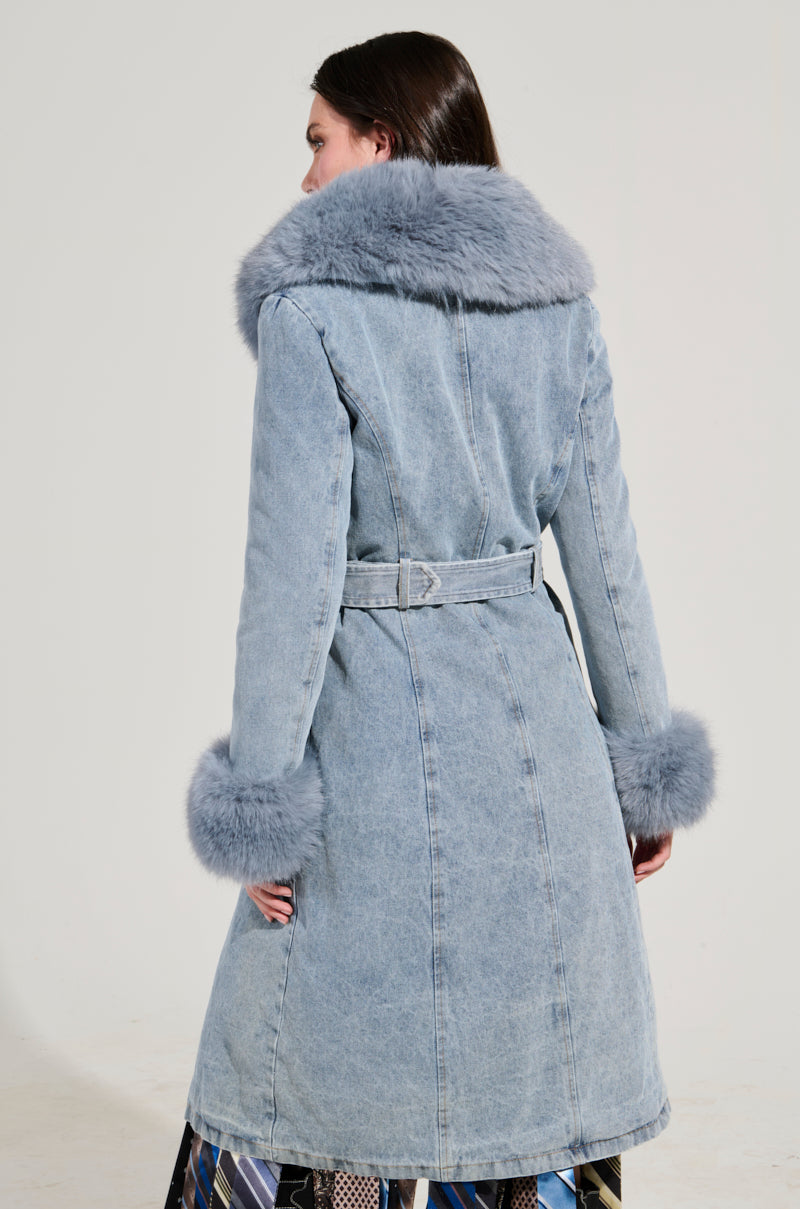 Someone is wearing the KAYA FUR TRIM TRENCH IN DENIM, a long light blue denim coat with a wide belt, large faux fur collar, and matching fur cuffs. They are facing away against a plain, light background.